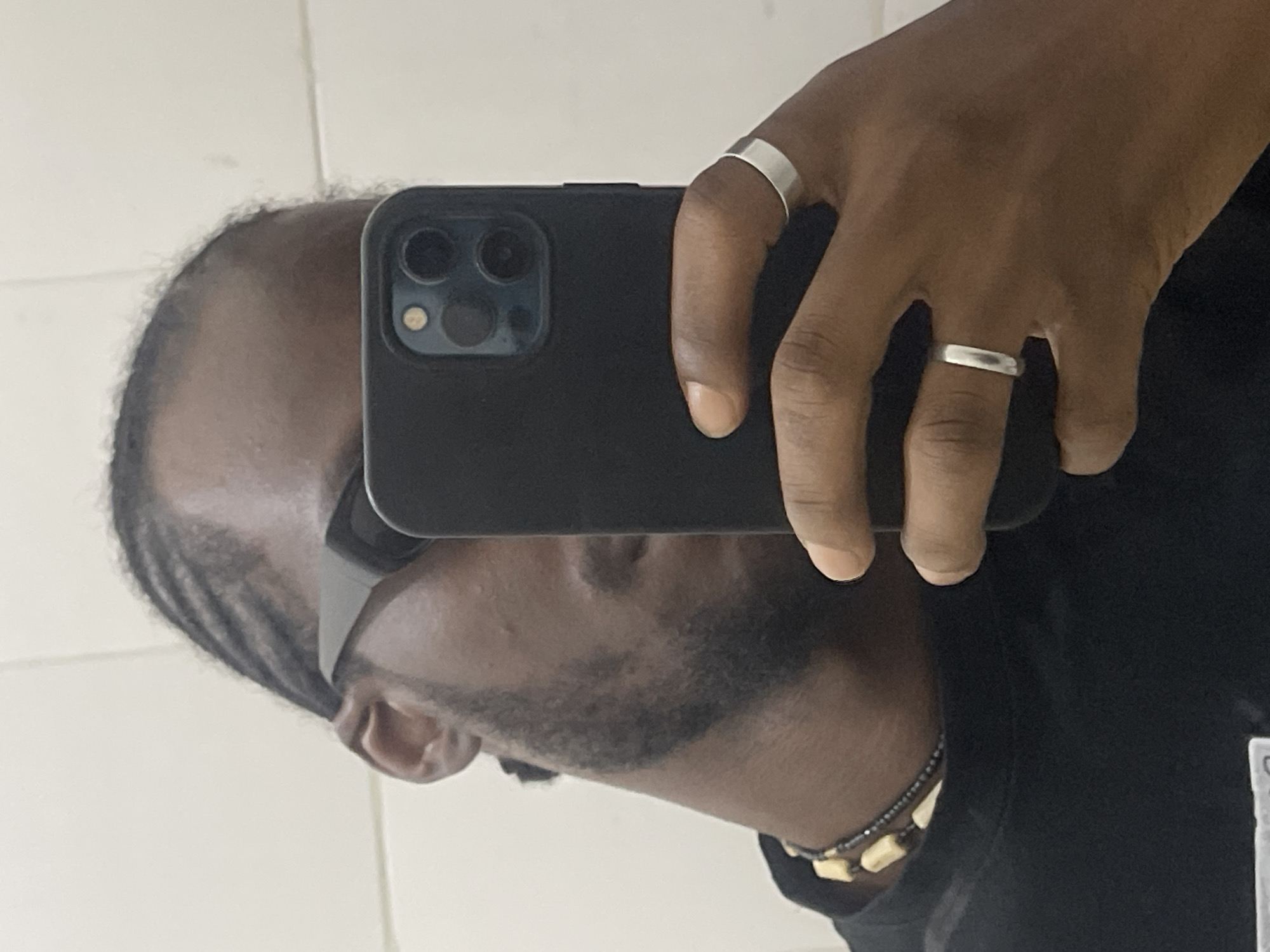 A person of dark complexion is depicted in a mirror selfie taken indoors in Jos, Nigeria. The subject exhibits braided hair (cornrows) on the visible side of the head and a trimmed beard along the cheek and jawline. Dark-framed sunglasses are worn on the head. The subject is dressed in a dark crew-neck t-shirt and a beaded necklace consisting of small dark beads and larger, light-colored rectangular beads. A black smartphone, featuring a triple-lens camera system, is held by the subject's left hand, which partially obscures the lower face. The left hand wears a wide silver-toned ring on its index finger. A narrower silver-toned ring is visible on the ring finger of the subject's right hand. The background is a plain, light-colored wall, likely tiled or paneled, indicating an interior location.