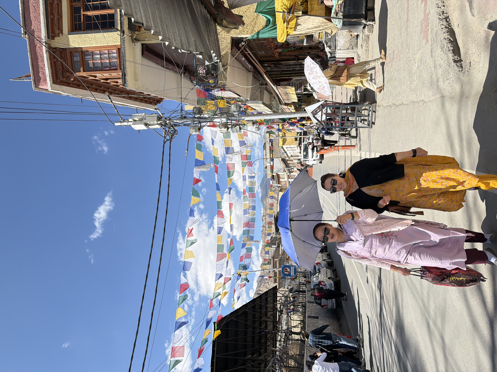 This is a full-shot, eye-level, outdoor photograph taken in Leh, India. The scene depicts a street scene with two women walking towards the camera under a blue sky adorned with prayer flags strung across utility wires. The woman on the left wears a light pink outfit and holds a colorful shopping bag. The woman on the right is dressed in a yellow patterned dress and a black cardigan, also holding a shopping bag. Both women are wearing sunglasses and appear to be conversing. A large blue umbrella is held over the woman on the left, casting a shadow. In the background, buildings line the street, displaying traditional Tibetan architecture with colorful trim and wooden windows. Several cars are parked along the side of the road, and other pedestrians are visible further down the street. The ground is paved with asphalt. The overall atmosphere is bright and sunny, suggesting a warm day in the mountainous region.