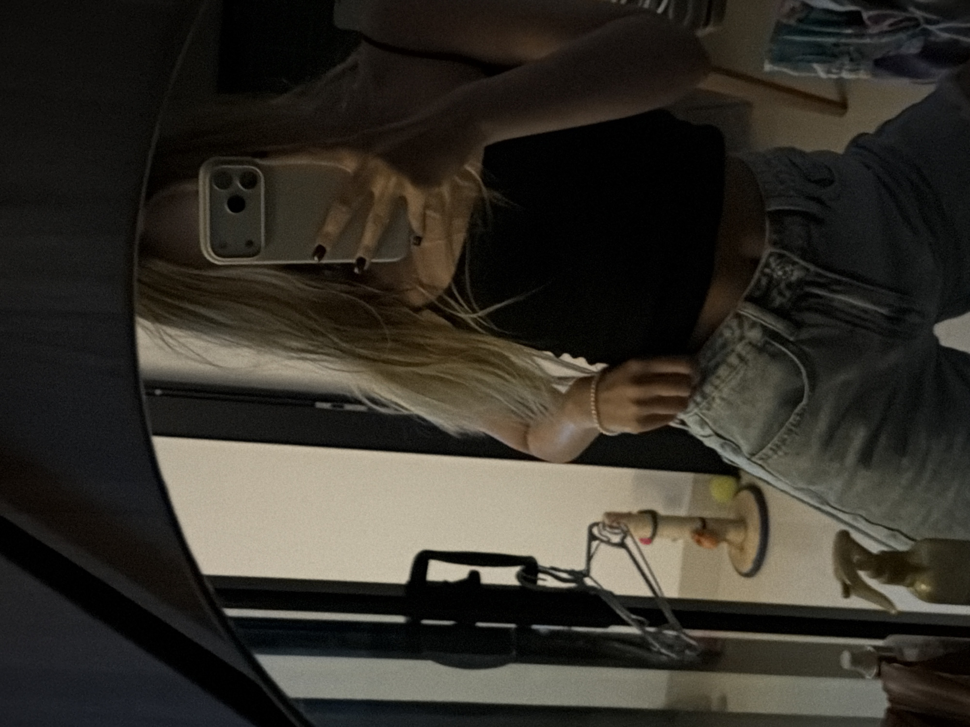 A person is taking a mirror selfie. They are wearing a black tank top and light-wash denim shorts. They have blonde hair and dark nail polish. In the background, a cat scratching post and a yellow ball are visible. A spray bottle is on the floor in the foreground. The scene appears to be indoors, possibly in an apartment or hotel room. The image does not provide enough information to determine the specific location in Da Nang, Vietnam.