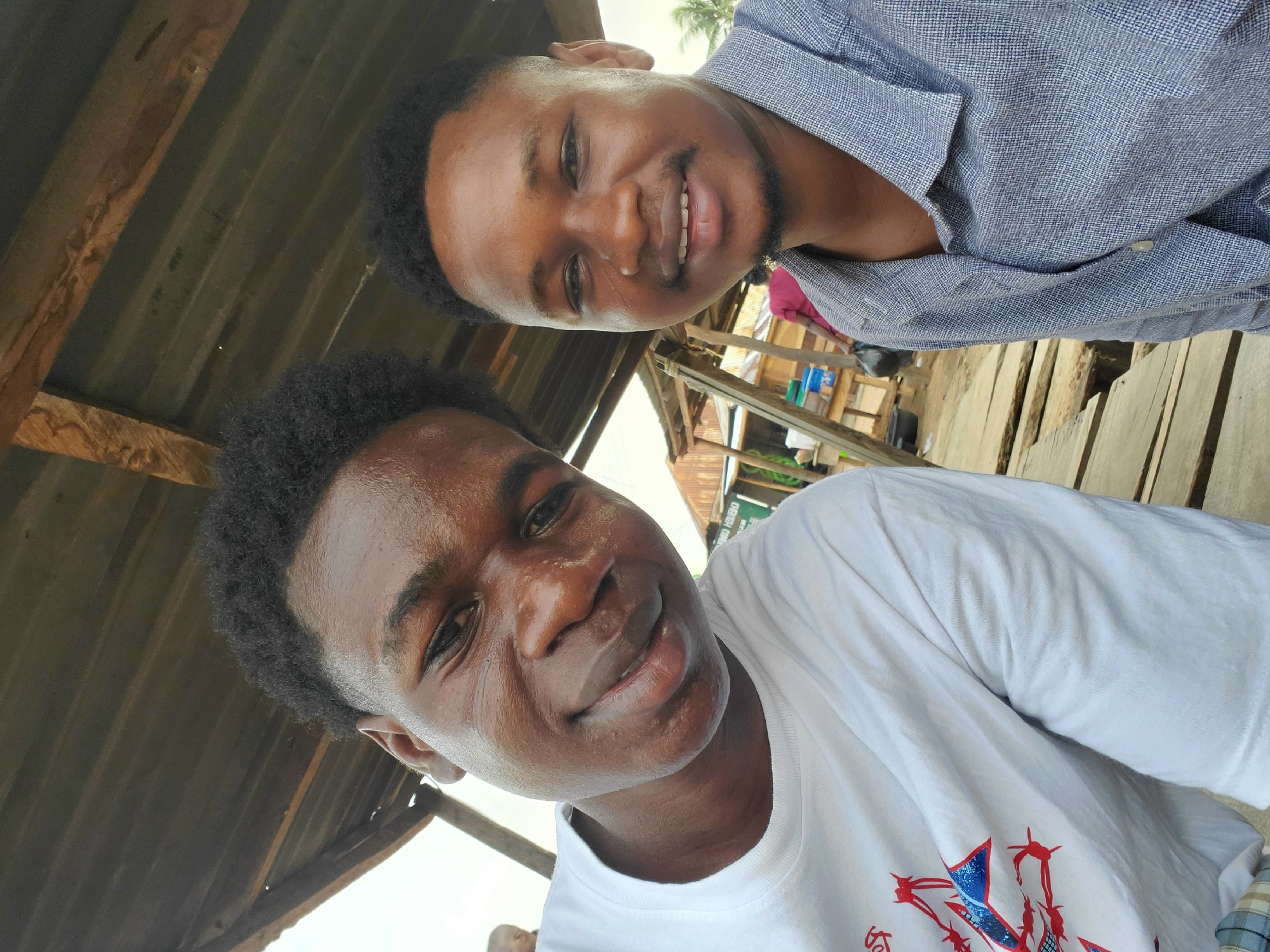 Two young men are featured in a close-up, slightly tilted selfie-style portrait, taken outdoors in what appears to be a semi-sheltered area. Both are smiling broadly and looking directly at the camera, conveying a friendly and cheerful mood.
The man in the foreground is on the right side of the frame. He has dark, curly hair and a clean-shaven face. He is wearing a white short-sleeved T-shirt with a partially visible graphic design on the chest, which includes elements of red and blue. His skin appears slightly dewy, perhaps from the weather or exertion.
Behind and slightly to the left of the foreground subject is another young man. He also has dark, short hair and a neatly trimmed beard/mustache. He is wearing a light blue or gray collared shirt with a subtle textured pattern.
The background suggests a casual, outdoor environment, consistent with areas found in Mararaba, Nigeria. Above the subjects, a dark, corrugated metal roof or ceiling is visible, supported by wooden beams, indicating a sheltered structure. Behind the man in the background, there are glimpses of wooden scaffolding or rustic structures, possibly part of market stalls or simple buildings. The lighting is bright and natural, typical of daytime. No discernible text is visible on clothing or in the background.
