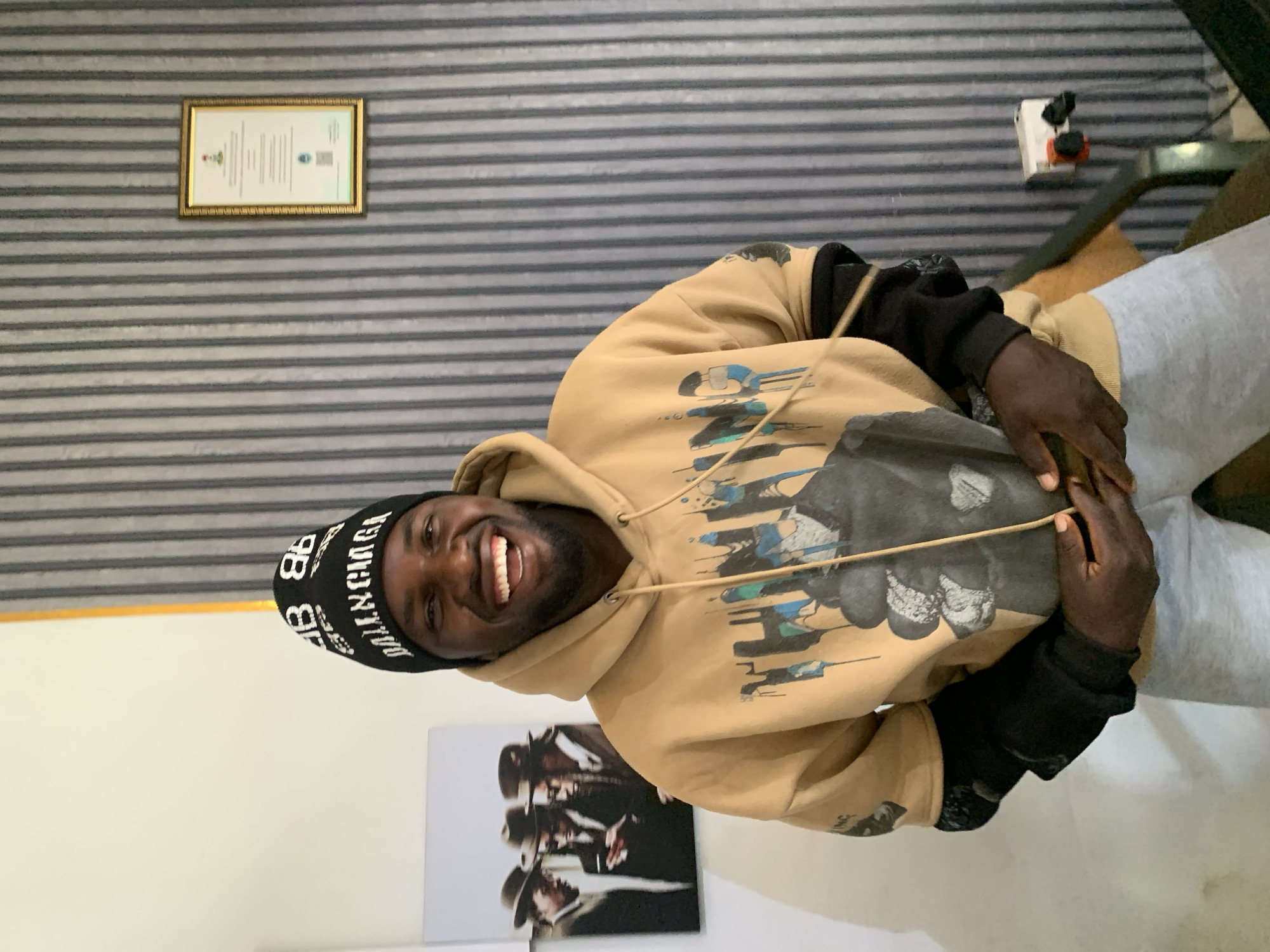 A man smiles at the camera, wearing a black Balenciaga beanie, a tan hoodie with a graphic design, and gray sweatpants. He is inside a room with a gray and white striped wall, a framed certificate on the wall, and a black and white art print on the lower wall. A power outlet is also visible. The location is Jos, Nigeria.