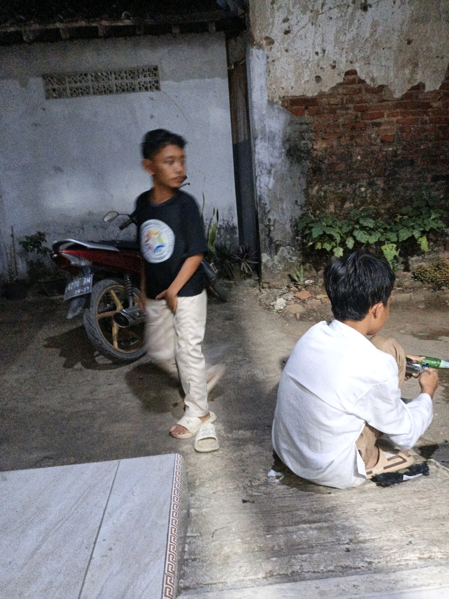 In Tanahbaya, Indonesia, two young males are present in an outdoor area at night. One boy, wearing a black t-shirt with a circular graphic and light-colored pants, is standing near a red motorcycle. He appears to be in motion, with his body turned away from the camera and his head looking to his right. The other boy, facing away from the camera and seated on the ground, wears a white long-sleeved shirt and tan shorts. He is holding a green and silver toy gun. The background features a weathered brick wall with patches of exposed brick and moss, and a concrete ground surface. There is also a tiled surface in the foreground.