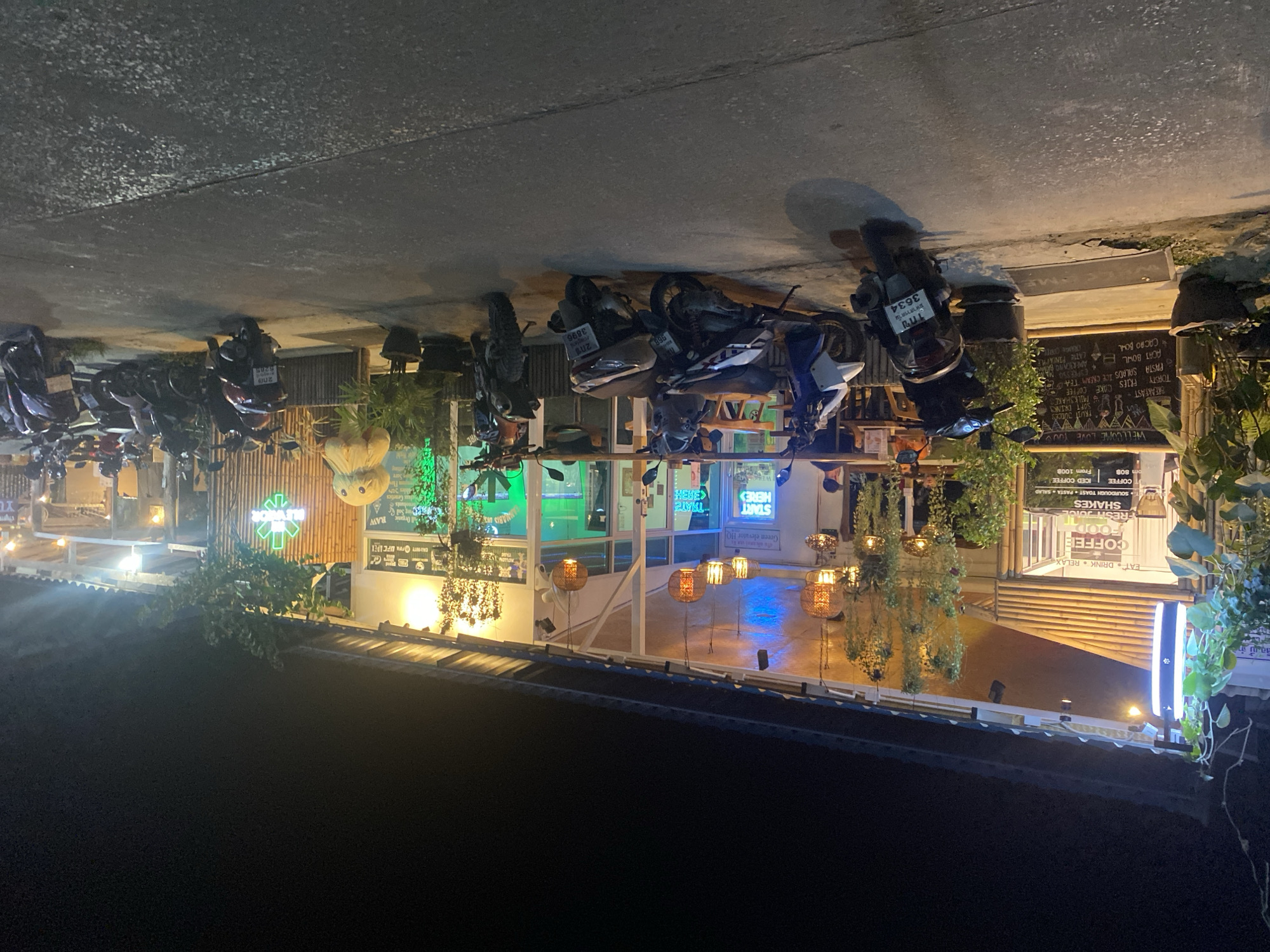 This is an image of a street-side establishment at night, likely a cafe or restaurant, with several motorcycles parked outside. The scene is illuminated by artificial lights, including neon signs and hanging lamps, suggesting it is after dark. The facade of the building appears to be made of bamboo and has large windows, revealing a brightly lit interior with tables and chairs.
Visible text on signs indicates a menu with items like "COFFEE," "FOOD," "FRESH JUICE," "SHAKES," "BREAKFAST," "PASTA," "SALADS," "ICE CREAM," and "CAKE." Some signs also feature slogans like "EAT DRINK RELAX" and "WELLCOME LOVE YOU." There are also Thai characters and license plates visible on the motorcycles, suggesting an Asian location. One sign displays "Green elevator HO" and an arrow pointing to "START HERE."
The overall atmosphere seems inviting and casual, possibly catering to a late-night crowd. The presence of many parked motorcycles implies that the establishment is popular and accessible by motorbike. The image is shot from a slightly elevated angle, looking down onto the street and the building. There are no people clearly visible in the foreground, but the interior suggests activity within. The weather appears to be clear, as there are no signs of rain or other adverse conditions.