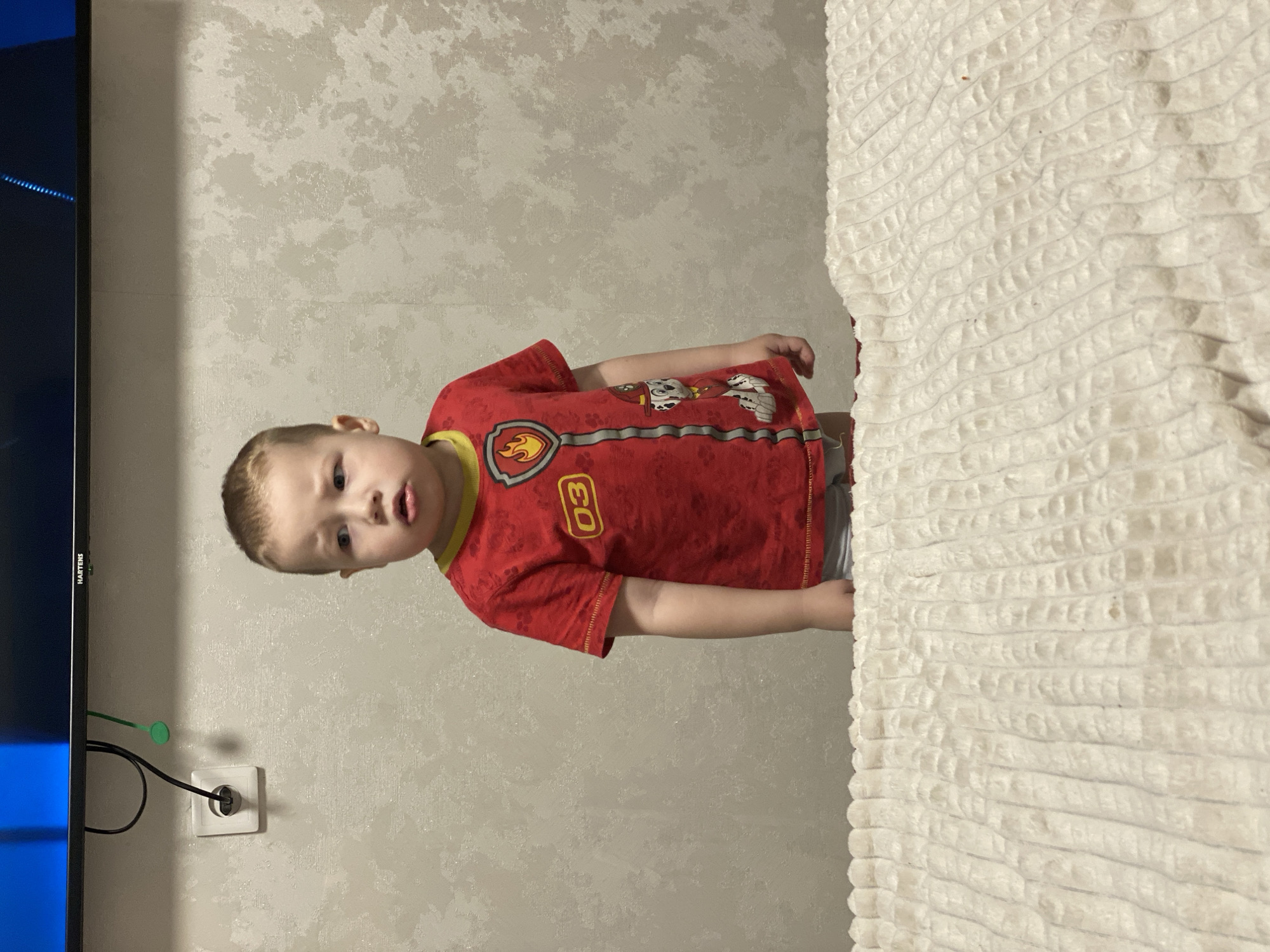 The image captures a young child, appearing to be a boy aged approximately 2-4 years, with fair skin and short light-colored hair, standing indoors. He is wearing a red short-sleeved t-shirt featuring a Paw Patrol character (Marshall), a yellow collar, and a yellow "03" badge with a flame emblem. Light gray undergarments are visible beneath the t-shirt. The child is positioned behind a textured, cream-colored blanket or comforter with a raised, ribbed pattern, which occupies the lower third of the frame. His head is turned slightly to the viewer's left, with eyebrows slightly furrowed and mouth slightly open, indicating an attentive or questioning expression. The background consists of a light-colored wall with a subtle, irregular pattern. Above the child's head, the lower portion of a black television screen, branded "HARTENS," is partially visible, displaying a blue image. Below the television, a white electrical outlet is mounted on the wall, with a black cable plugged into it and a small green cable organizer attached to the TV bezel. The scene is located in the city of Sochi, Russia.