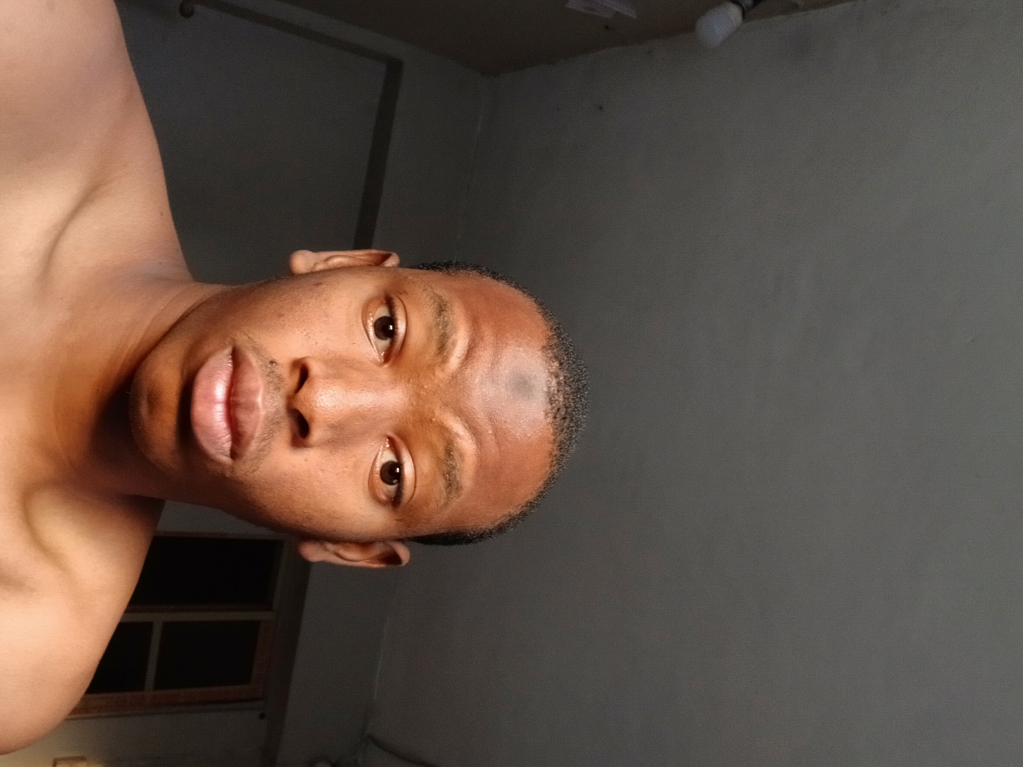 An intimate, sideways-oriented portrait features a young man with dark skin and closely cropped dark hair, likely taken in Lagos, Nigeria. He is shirtless, displaying his bare shoulder and upper chest. The man gazes directly into the camera with wide, expressive brown eyes and a slightly open mouth, revealing a hint of his teeth. His skin appears somewhat reflective, possibly due to lighting or perspiration.
The background consists primarily of a plain, dark gray wall, suggesting an indoor setting. In the upper right corner, a bare light bulb hangs, serving as the primary artificial light source, which casts soft shadows on the subject's face and body. To the lower left, a dark, rectangular structure with lighter framing, possibly a window or a partition, is partially visible. The dim lighting conditions suggest the photo was taken either at night or in a dimly lit room. No text is visible in the image.