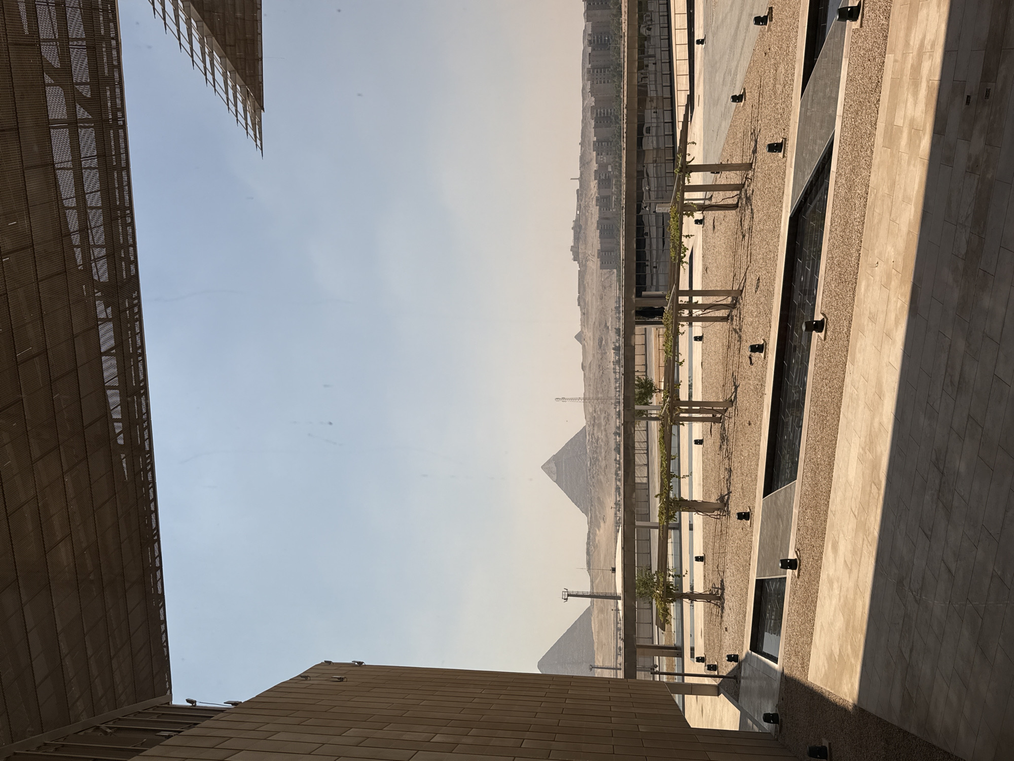 The image depicts a modern architectural structure in the foreground, with a clear view of the iconic pyramids of Giza in the distance, suggesting the location is Kafrat Nassar, Egypt. The foreground features a paved area with gravel beds and shallow water features, accented by strategically placed black spotlights. Several slender trees with overhead trellises are spaced throughout the gravel areas. The left side of the image is dominated by a portion of a tan, textured building, extending upwards and partially obscuring the sky. Above the building and the outdoor area, a metallic, perforated canopy stretches across the top of the frame, casting subtle shadows.
In the background, the vast expanse of the desert landscape is visible. Prominently, the Great Pyramid of Giza and the Pyramid of Khafre stand as distinct triangular shapes against the hazy sky. Between the modern foreground and the ancient pyramids, a low-rise urban development with numerous buildings stretches horizontally, interspersed with some greenery. The sky is a pale, cloudless blue, indicating a clear and likely warm day. There are no people visible in the scene. The overall impression is one of juxtaposition between contemporary architecture and the ancient wonders of Egypt.