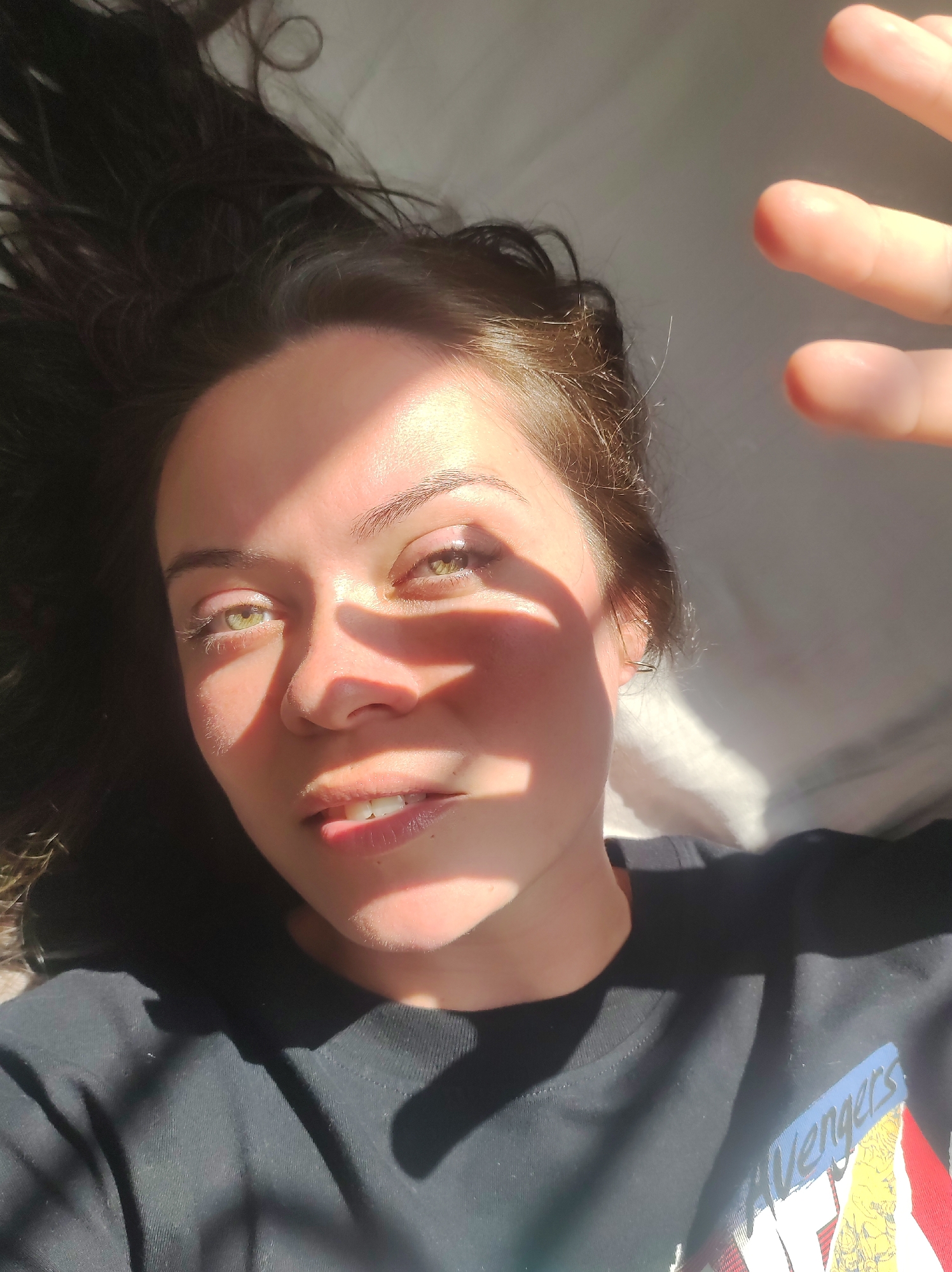 A young woman is lying on her back, with her head tilted to the right. She has long, dark hair that is spread out around her. Sunlight is falling on her face, creating shadows. She is wearing a black t-shirt with the word "Avengers" on it. The background appears to be a white sheet or blanket. The scene is likely indoors, possibly in a bedroom. The woman's eyes are open and she is looking up towards the camera. Her right hand is raised, obscuring part of her face and the sun. The image is a close-up shot, focusing on the woman's face and upper body. There is no information available about the specific location in the city of Chaikovsky, Russia, or any other details about the setting.
