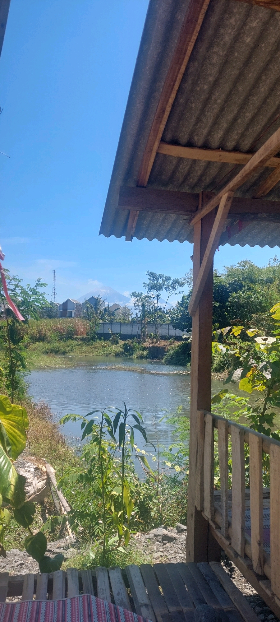 The image shows a view from under a rustic wooden shelter with a corrugated metal roof, looking out towards a body of water. The shelter's wooden posts and railing are visible in the foreground. Beyond the shelter, a calm body of water reflects the bright blue sky. On the opposite bank of the water, there is a line of vegetation, followed by houses and a telecommunications tower. In the far distance, a prominent, large mountain with a conical shape is visible, possibly Mount Bromo, a well-known volcano near Lumajang, Indonesia. The overall scene depicts a tranquil, outdoor setting with a natural landscape and a hint of human habitation.