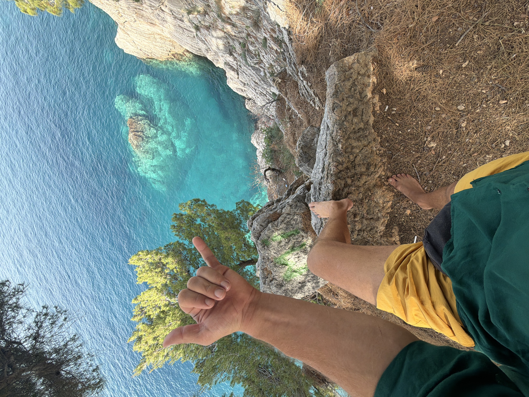A low-angle, rotated perspective shows a man standing on a cliff edge in Petrovac centar, Montenegro. He is barefoot and wears mustard-colored shorts with a dark green shirt over them. His right arm is extended, hand forming a "shaka" sign, with the little finger and thumb extended. The edge of the cliff is composed of weathered rock and pine needles. Below, the Adriatic Sea is turquoise, revealing rocky underwater formations. The city is not visible in this image. The cliff edge has a small cluster of trees growing over the side.