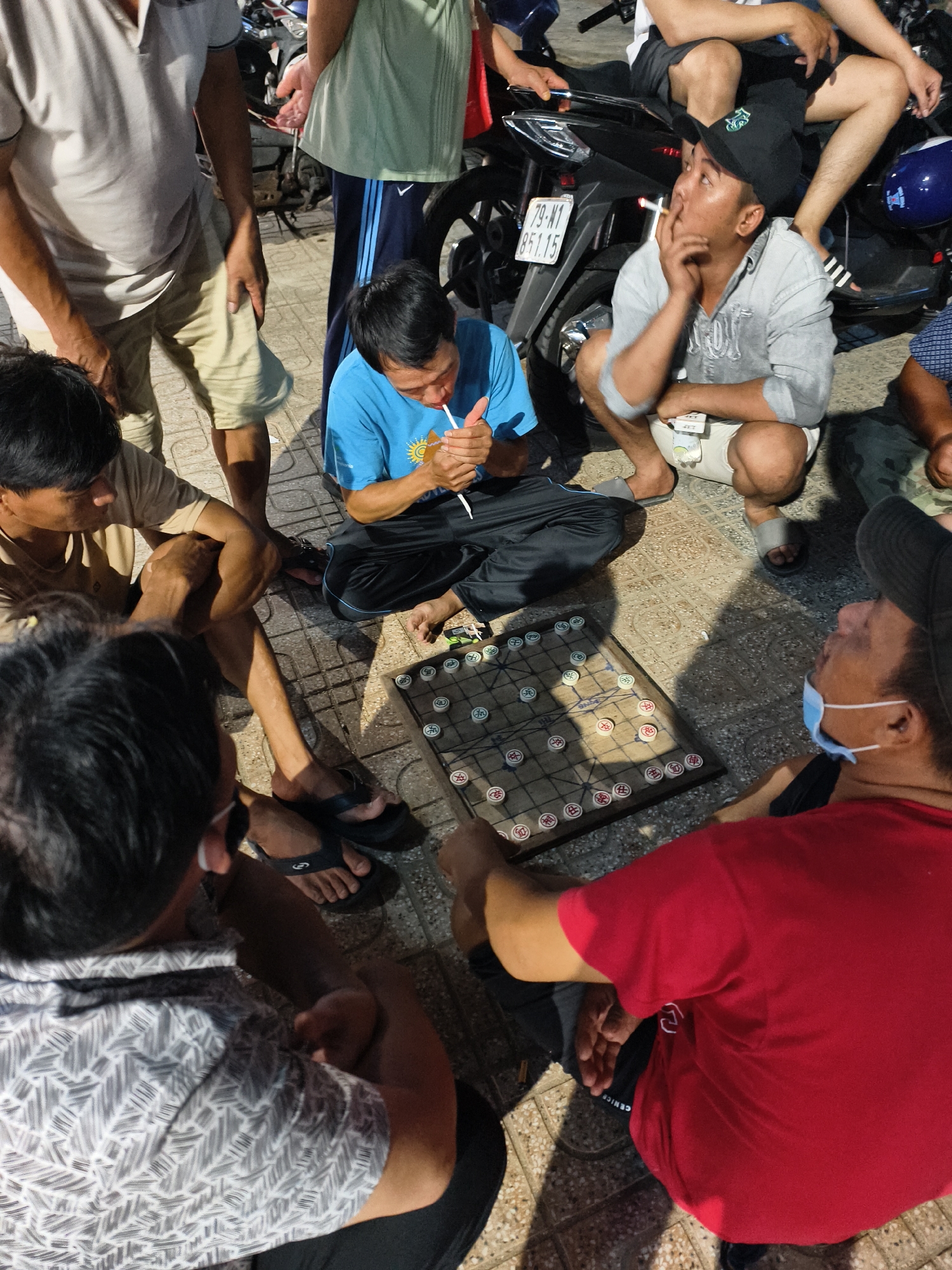In a street scene in the city of Unknown, Unknown, a group of men are gathered around a chess board on the ground. One man in a blue t-shirt is lighting a cigarette with a lighter. Another man, wearing a grey shirt and black cap, is smoking a cigarette and squatting. A man in a red t-shirt is wearing a face mask. There is a motorcycle parked nearby. The men are dressed in casual clothing. The ground is tiled with a geometric pattern.