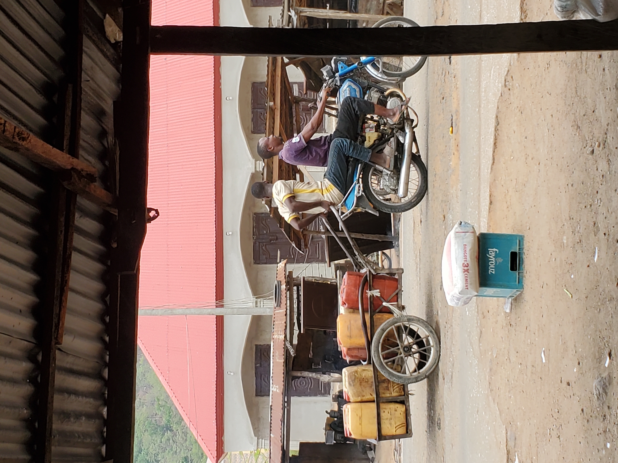 In Mararaba, Nigeria, a partially obstructed view of a street scene reveals two men on a motorcycle. One man, wearing a purple shirt and dark pants, appears to be working on the motorcycle's engine. The other man, in a yellow and white striped shirt, sits behind him. The motorcycle is modified to pull a cart filled with yellow jerrycans. A blue crate and a bag are near the cart. The buildings behind the men have red roofs and multiple windows. The street appears to be dirt or gravel. A corrugated metal structure obstructs part of the view.