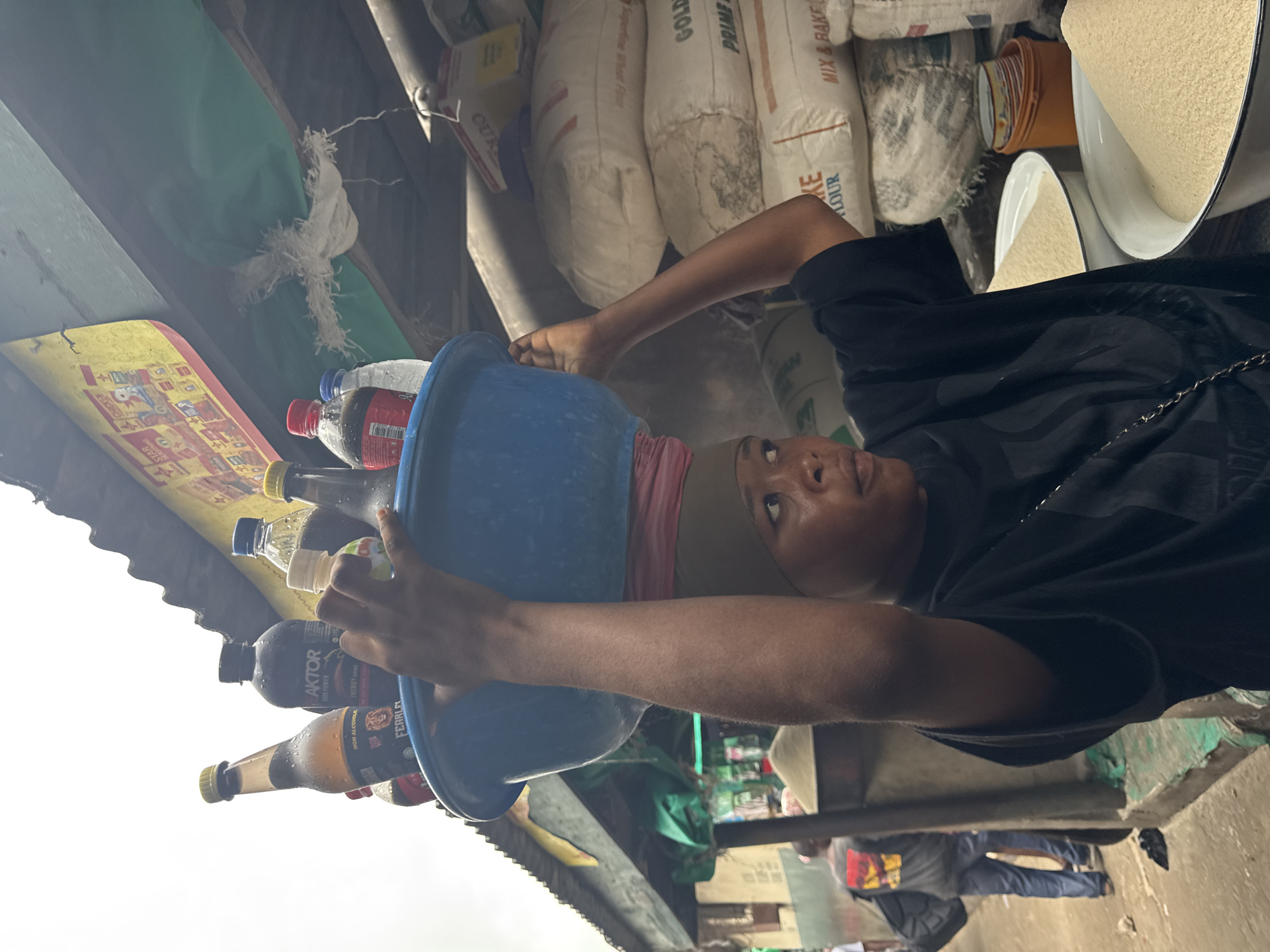 In an outdoor market scene in Lagos, Nigeria, a young woman balances a blue basin filled with bottles on her head. The bottles appear to contain beverages, including one labeled "AKTOR POWER DRINK" and another that is amber-colored with a yellow cap. She wears a dark t-shirt and a headscarf. The background shows stacks of large white sacks, likely containing flour or grains, and a green tarpaulin overhead. In the distance, a person in a grey shirt and blue jeans walks away from the camera. The ground is a rough concrete surface.