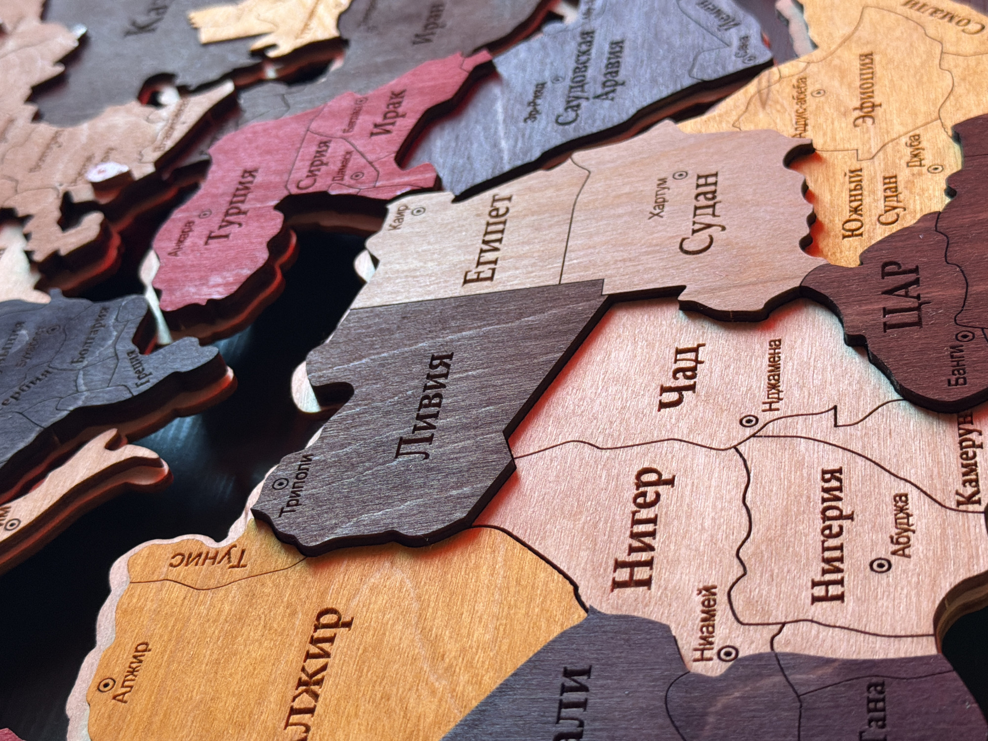 This is a close-up, top-down shot of a wooden world map puzzle. The puzzle pieces are cut out in the shapes of countries and are engraved with their names in Cyrillic script. The wood grain is visible on each piece, and some pieces have a reddish-brown or darker wood tone, while others are a lighter, natural wood color. The map focuses on Europe, the Middle East, and Africa, with countries like Turkey, Syria, Iraq, Egypt, Sudan, Libya, Niger, Nigeria, Ethiopia, and others clearly labeled. Small circles are engraved within some countries, likely indicating capital cities. The overall impression is that of an educational and decorative item, possibly a handicraft. There are no people visible in the image. The lighting suggests an indoor setting, with shadows cast by the raised puzzle pieces, indicating the light source is likely from the side and slightly above. The context of Orenburg, Russia is noted, but not directly evident from the visual information in the image itself.