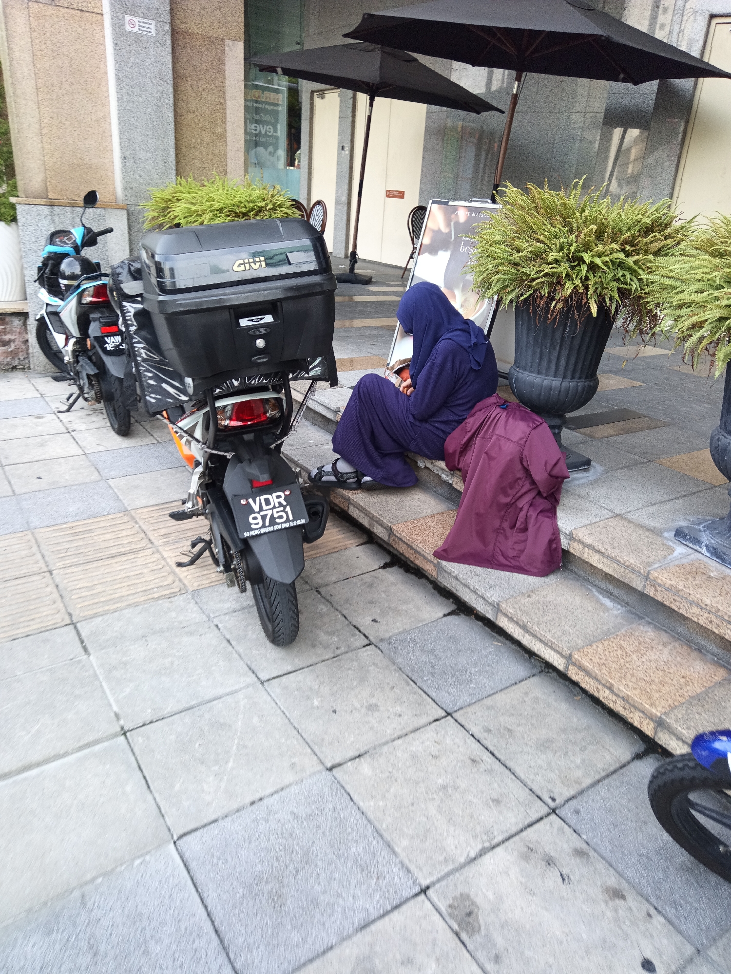 The image depicts an outdoor scene on a paved sidewalk in Kuala Lumpur, Malaysia. Two motorcycles are visible, one parked to the left and partially obscured by a potted plant, and another more prominently displayed in the foreground. The foreground motorcycle is black with orange accents, features a large black Givi top case, and has a license plate displaying "VDR 9751". A person wearing a purple hijab and a matching purple robe is seated on the steps to the right of the motorcycle, with a dark red jacket draped over the step beside them. The background shows the exterior of a building with granite cladding, large black umbrellas, and more potted plants. The lighting suggests an overcast day.