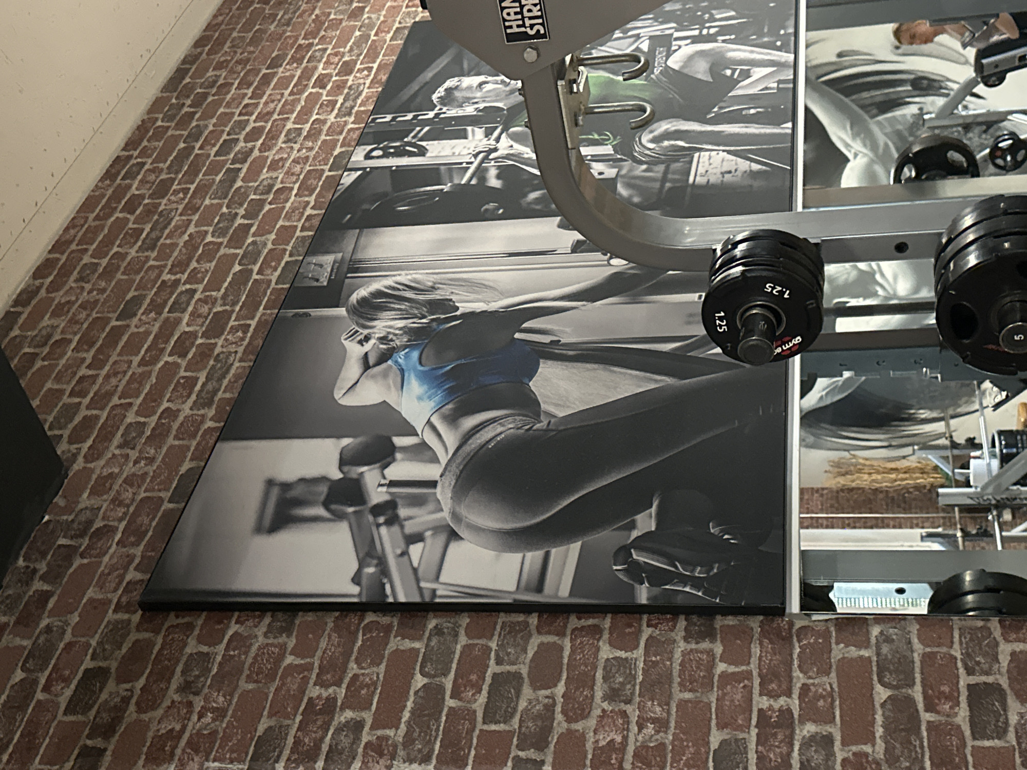 The image shows a section of a room with a brick floor and partial view of a wall. Affixed to the wall are two large, rectangular panels depicting scenes of fitness activities in black and white. In the foreground, a Hammer Strength brand weightlifting machine is visible. The panels, placed side-by-side, feature athletes in workout poses, one appears to be performing a squat. A set of weights are stacked on the weightlifting machine in the foreground. The image was taken in Brunnthal, Germany.