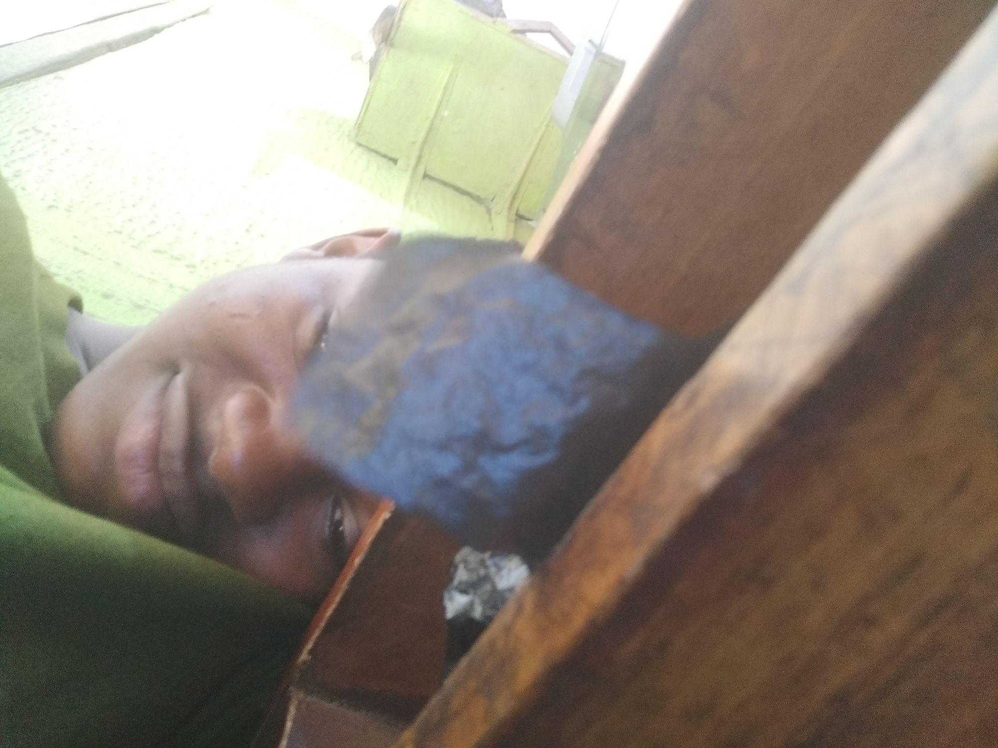 A low-angle close-up shot features a person lying down, partially visible in the lower left corner, wearing a green garment. A wooden structure dominates the right side of the frame, out of focus due to proximity. A dark, rectangular object with a textured surface is positioned near the center, possibly obscuring part of the wooden structure. In the background, an out-of-focus green object, potentially a cabinet or storage unit, is visible. The scene seems to be indoors. The location is stated to be Shangisha, Nigeria, but there are no visual cues in the image to confirm this.