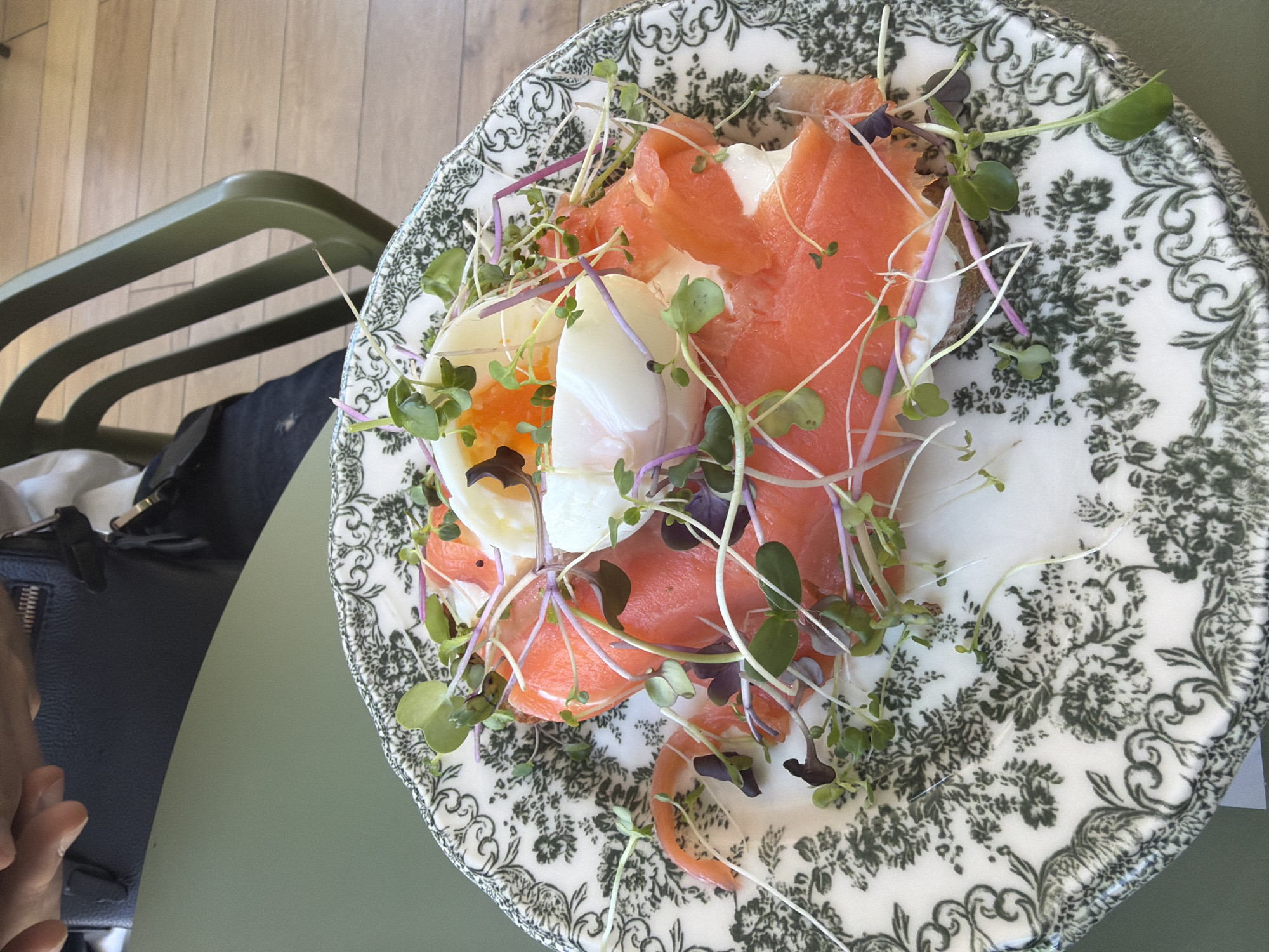 A high-angle shot depicts a plate of food, likely in Germasogeia Municipality, Cyprus, based on the general aesthetic and potential cafe/restaurant scene. The plate has a white base with a dark green floral pattern around the edge. The food consists of salmon slices, a poached egg with a visible yolk, and sprouts or small greens. The food appears to be placed on a piece of bread. The plate sits on a green table next to a black purse/bag. Part of a green chair and a wooden floor are visible in the background. The edge of a person's hand is visible in the bottom left corner.