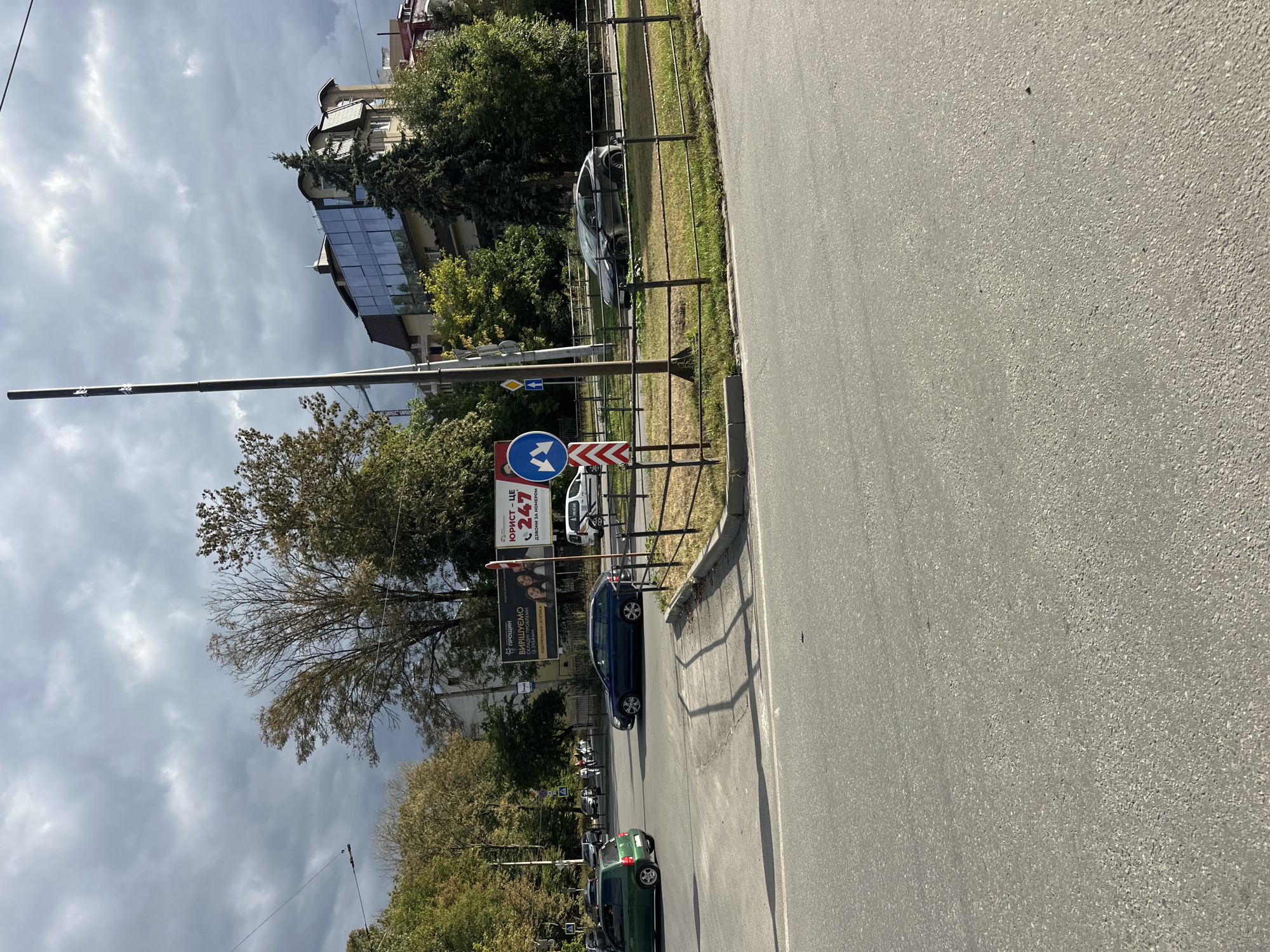 The image depicts a street scene in Ternopil, Ukraine, on an overcast day. In the foreground, a paved road with lane markings is visible. A traffic island with a metal fence separates lanes of traffic. Several cars are present on the road: a green hatchback, a dark blue sedan, and a dark grey hatchback, which appears to be a Tesla.
A tall, slender lamppost stands near the traffic island. Traffic signs are visible, including one with blue arrows indicating a lane split for left or straight turns, and a diamond-shaped yellow sign with a black arrow.
In the background, trees with green and autumn foliage line the street. A building with a modern glass facade and a curved roof is visible behind the trees. A large billboard advertising a legal service with the phone number "247" is also present, along with images of people. Other buildings, some with pitched roofs, are partially visible. The sky is heavily clouded with a mix of grey and lighter patches, suggesting potential rain. There are no people clearly identifiable as subjects of interest within the frame.