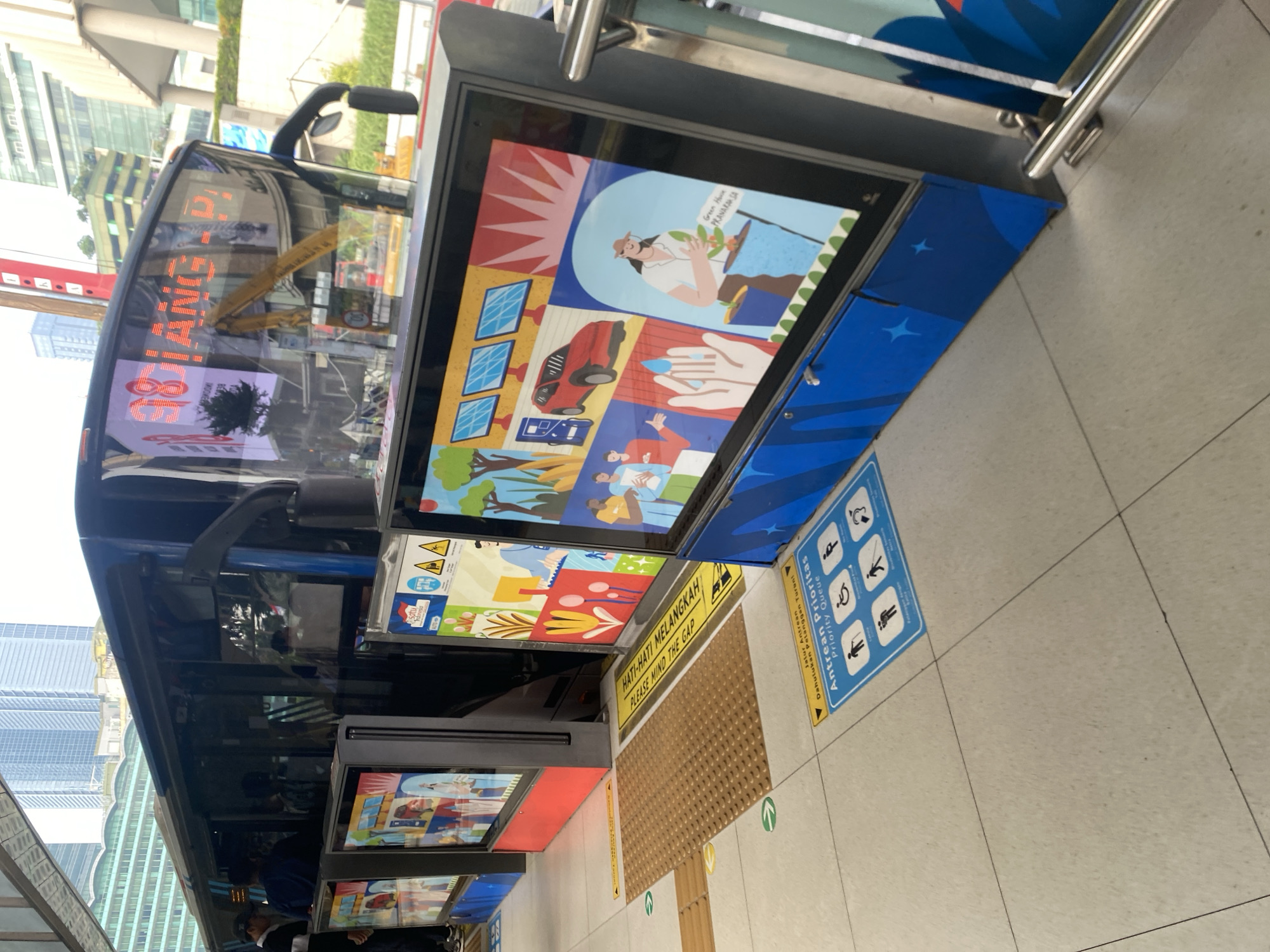 The image captures a bus stop in Central Jakarta, Indonesia. A bus with a digital display showing "98 DIANG. P.!" is parked at the station. In front of the bus are several digital screens displaying colorful graphics related to environmental themes, such as solar panels, electric cars, water conservation, and gardening. On the ground, there are signs for a "Priority Queue" for passengers needing special assistance, including the elderly, disabled individuals, and pregnant women. The floor is tiled, with yellow tactile paving indicating the edge of the platform. The background shows modern city buildings, suggesting an urban environment. A metal handrail is visible on the right side of the frame.