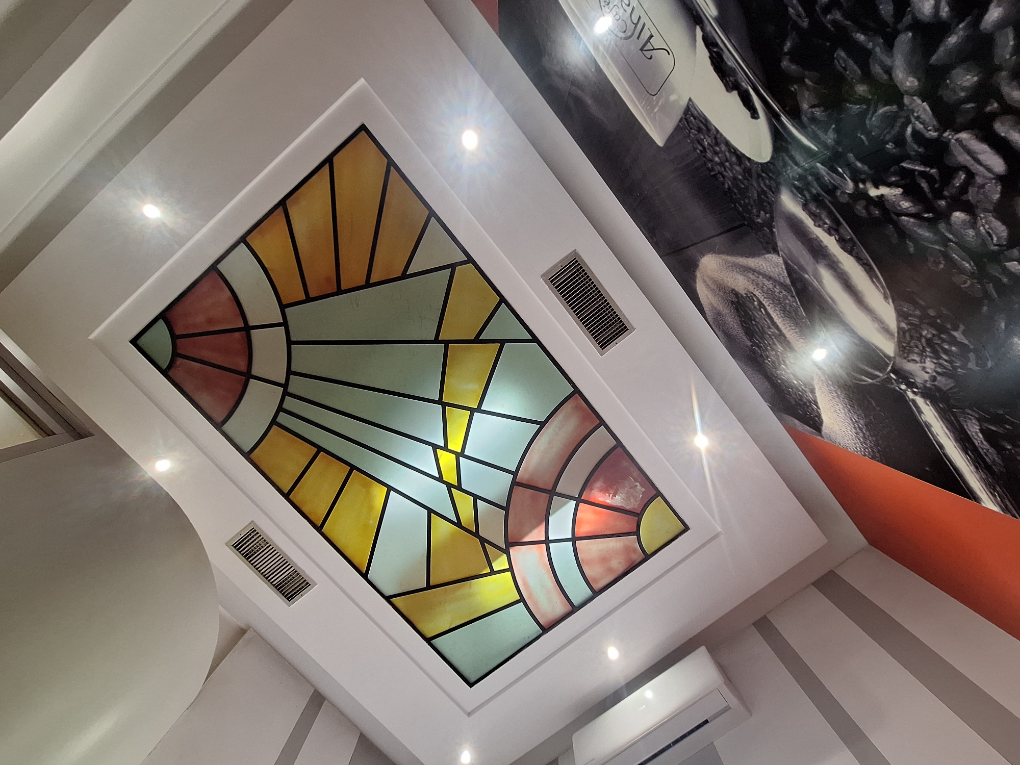 This interior image, taken in Casablanca, Morocco, captures an upward-angled view of a ceiling and an adjacent wall within what appears to be a commercial establishment, such as a café or lounge.
The ceiling is predominantly white, featuring multiple architectural levels and straight lines. A central, rectangular stained glass panel is embedded within a white recess. This panel exhibits an abstract, geometric design with segments colored in yellow, light blue/teal, and reddish-brown/orange, all outlined by black lines. The panel is illuminated from behind, creating a luminous effect. Five bright, circular recessed spotlights are visible on the white ceiling, casting starburst lens flares. Two rectangular air vents with horizontal grates are integrated into the ceiling, one positioned above the stained glass panel and another below it. In the far upper-left corner of the image, a smooth, curved white architectural element is present, and to its left, a white wall-mounted air conditioning unit is partially visible.
The adjacent wall, visible in the lower right portion of the image, is covered by a large mural or wallpaper. The mural features a coffee-themed design in a largely monochromatic palette of greys, blacks, and whites, with subtle brown tones from the depicted elements. A white coffee cup and saucer are shown on the right, with partial text "Café Alha..." visible on the cup. A metal scoop filled with coffee beans is depicted in the foreground, surrounded by scattered roasted coffee beans. An angular, bright orange panel or wall section acts as a contrasting element between the white ceiling structure and the coffee mural on the right side.
No individuals, actions, or direct interactions are visible within the scene.