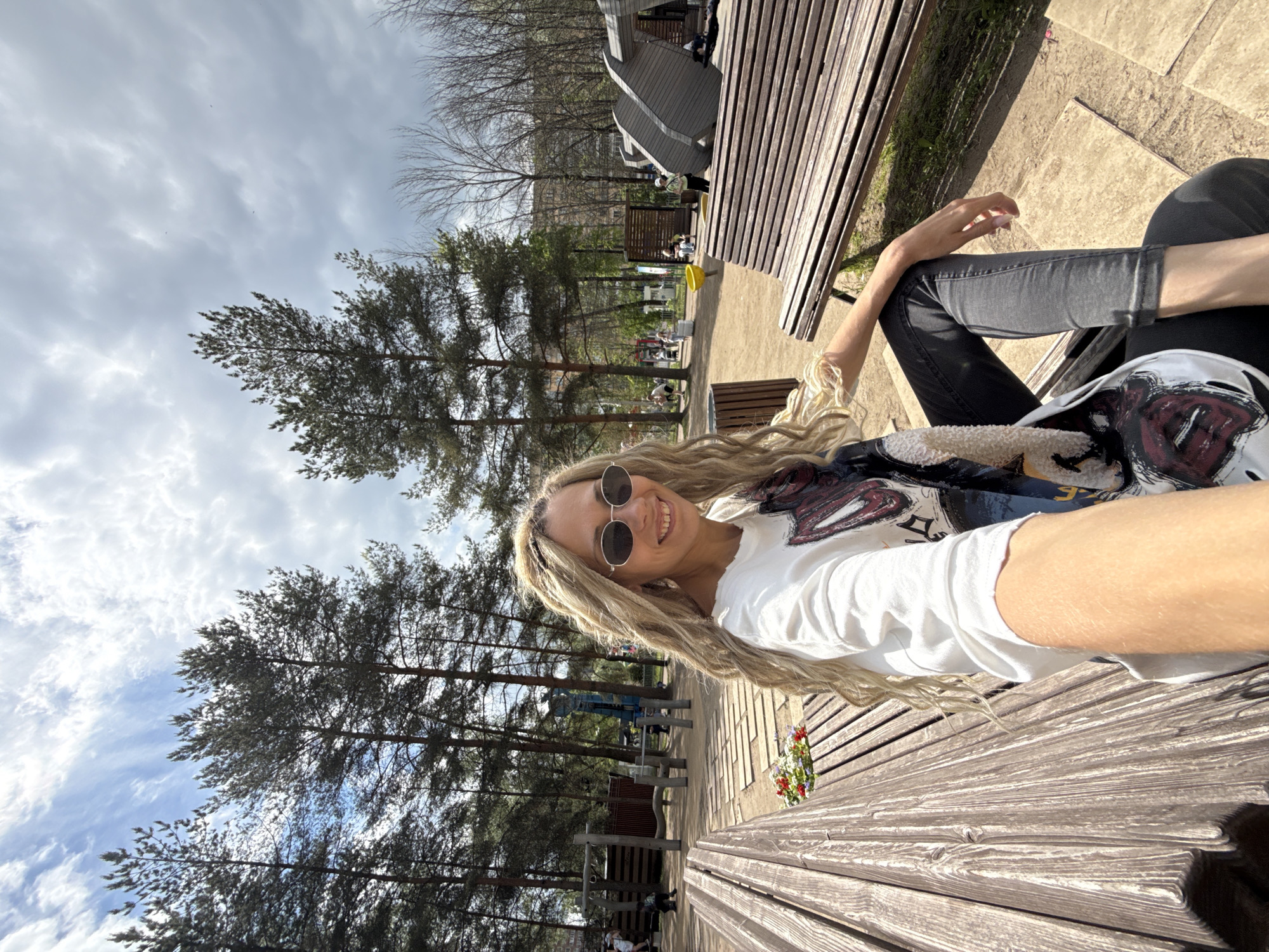 A smiling woman with long, wavy blonde hair and sunglasses takes a selfie, sitting casually on a wooden park bench. She is wearing a white t-shirt adorned with an abstract graphic design and dark trousers. Her left arm is visible, holding the camera for the shot.
The scene is an outdoor park in Шебунино, Russia, under a bright, partly cloudy sky with visible blue patches. Tall pine trees are prominent throughout the background, along with some bare deciduous trees in the distance. The park features include dirt paths, paved areas, and various wooden structures such as additional benches and what appear to be playground or exercise equipment. A small, colorful flower arrangement in a pot is visible on the bench to the woman's right. The overall impression is a pleasant and relaxed daytime spent in a public recreational area.
