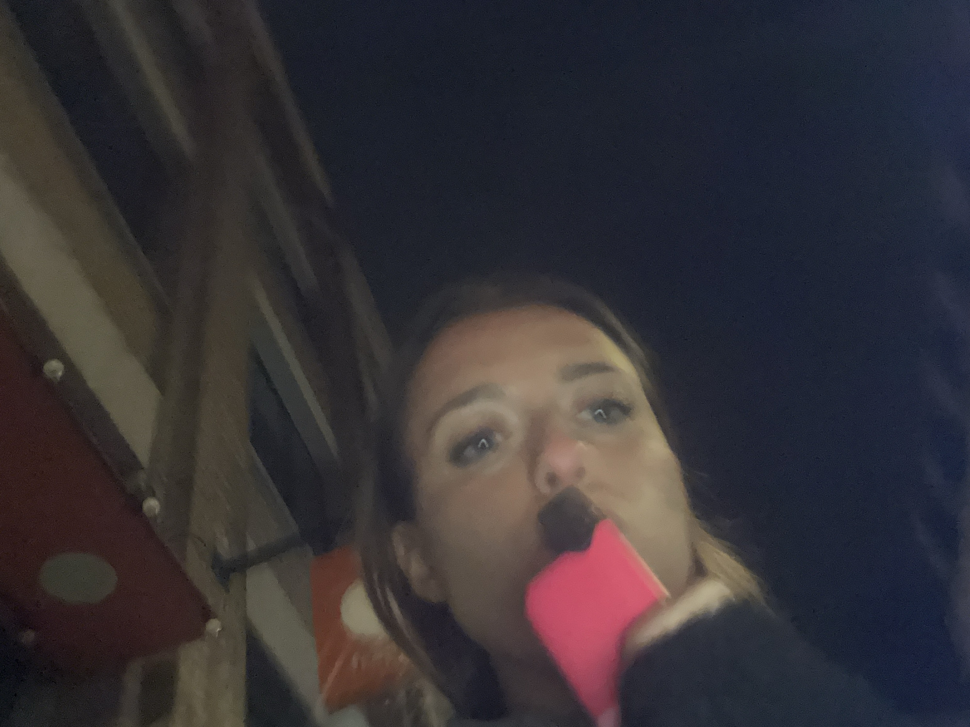 A low-angle, close-up photograph captures a person holding a pink electronic cigarette to their mouth. The individual has light skin, brown hair, and light-colored eyes. To the left, out of focus, is a red awning-like structure with circular indentations. Above the person, the dark sky creates the backdrop.
