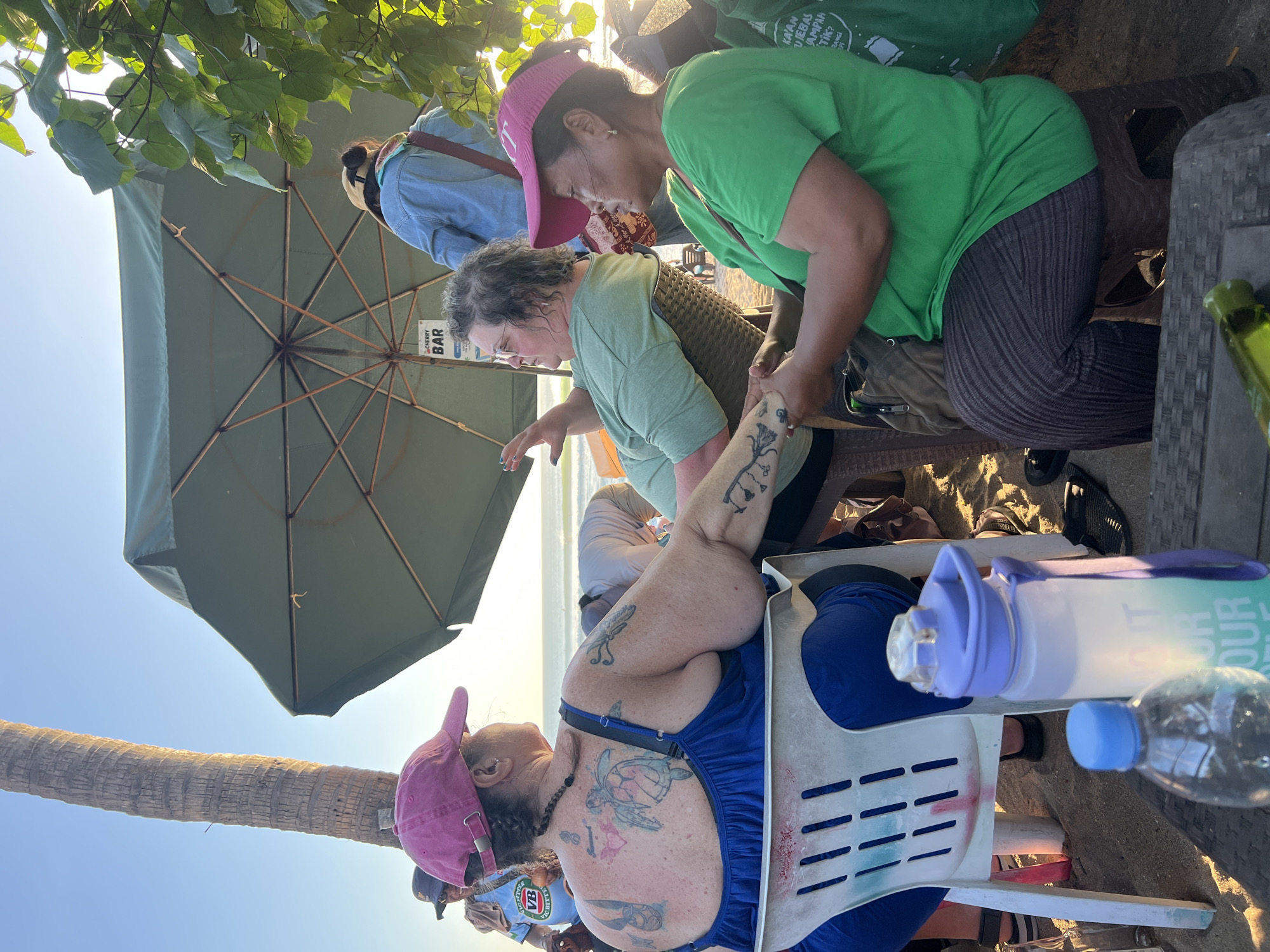 At a beachside establishment in Kuta, Indonesia, a group of people are gathered under a large green umbrella. The sand is visible in the foreground and around the seating area. Two women are in the foreground, seated and engaged in conversation. The woman on the left, wearing a blue dress and a pink baseball cap, has numerous tattoos visible on her back and arms. She is sitting in a white plastic chair. To her right, a woman in a green t-shirt and dark striped pants is seated on a brown plastic stool, holding hands with the woman in blue. Another woman in a green t-shirt is visible in the background, as is a person wearing a denim jacket. A sign that reads "CHERRY BAR" is attached to the umbrella. In the foreground, two water bottles are placed on a table. The overall lighting suggests it is late afternoon or early evening, with the sun casting a warm glow. The ocean and beach are visible in the background, indicating a coastal setting.