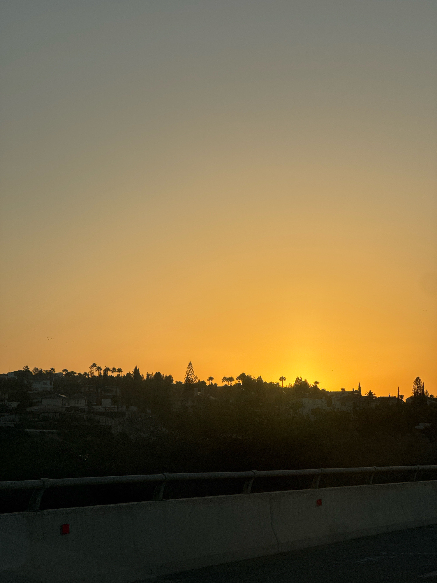 This image captures a sunset over Germasogeia Municipality, Cyprus. The foreground features a concrete barrier with reflective markers, likely part of a road or overpass. Beyond the barrier, silhouetted against the vibrant orange and yellow sky, is a hillside populated with houses and trees, including several palm trees. The sun is low on the horizon, casting a warm glow and creating a serene, twilight atmosphere. The sky transitions from a soft gray at the top to a rich orange near the horizon, with a few faint birds visible in the distance.