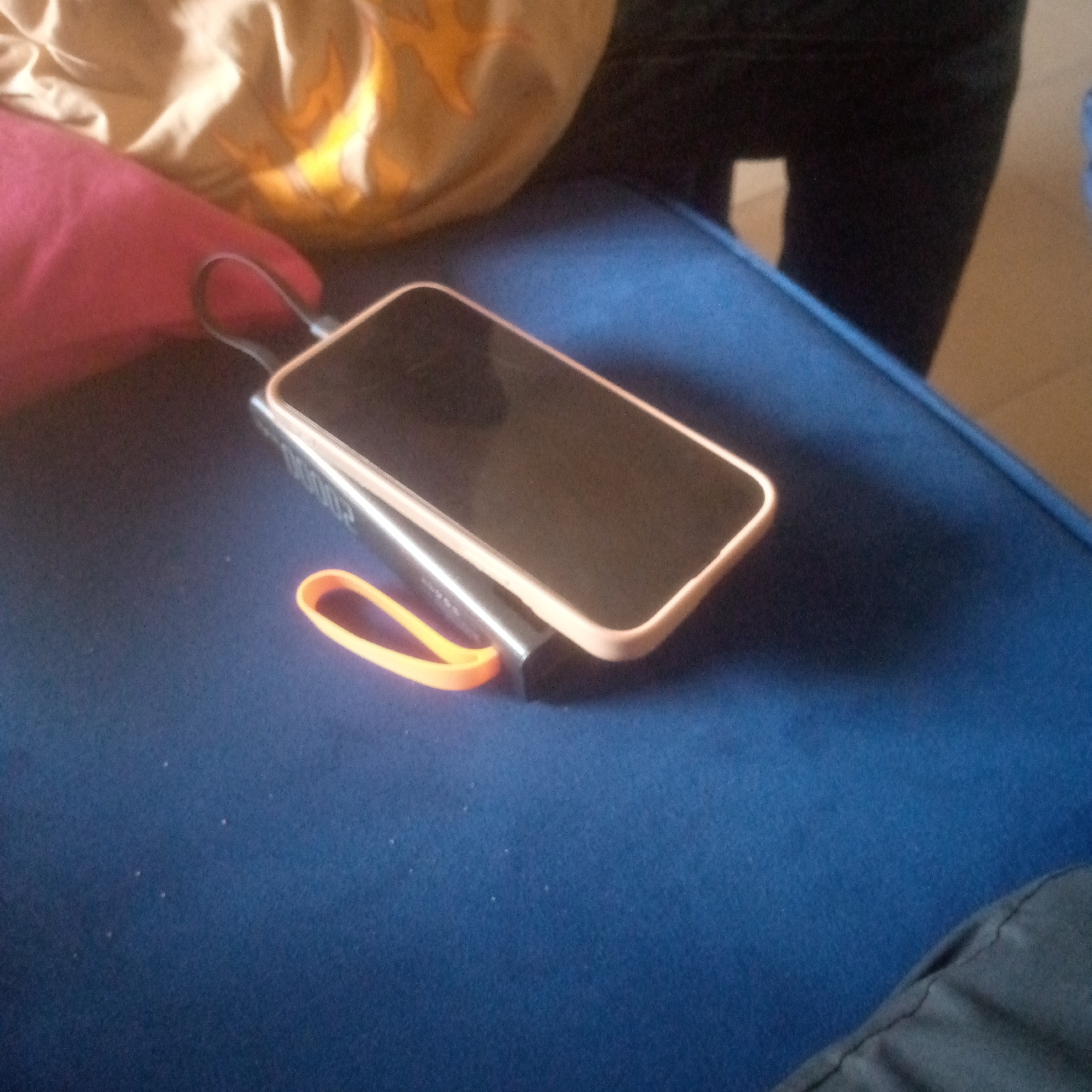 The image depicts an indoor scene at an unknown location, with no identifiable individuals present. The central subjects are a smartphone and a power bank resting on a dark blue upholstered surface, consistent with a couch or chair cushion. The smartphone, enclosed in a light pinkish-beige case, is positioned face-up with a dark, inactive screen. It rests directly on top of a black power bank, which displays "20000" in white text on its upper side. A silver charging cable connects the smartphone to the power bank. Adjacent to the connected devices is a bright orange silicone loop or strap. In the upper left quadrant, a crumpled light beige fabric with subtle orange and yellow patterns is visible. The right side of the background reveals dark fabric, likely clothing, against a lighter, tan-colored surface, potentially a floor. The scene is illuminated by ambient light.