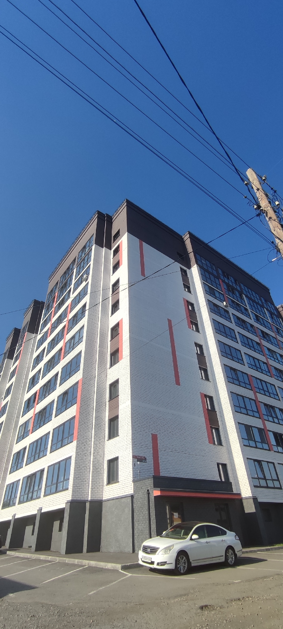 This is a vertical, eye-level, outdoor, medium shot of a modern apartment building in Novoaltaysk, Russia. The building has a white brick facade with vertical red accents and a dark grey top. The windows are tinted. A white sedan is parked in front of the building on a paved lot. The sky is clear and blue, and there are utility wires visible in the upper part of the frame.