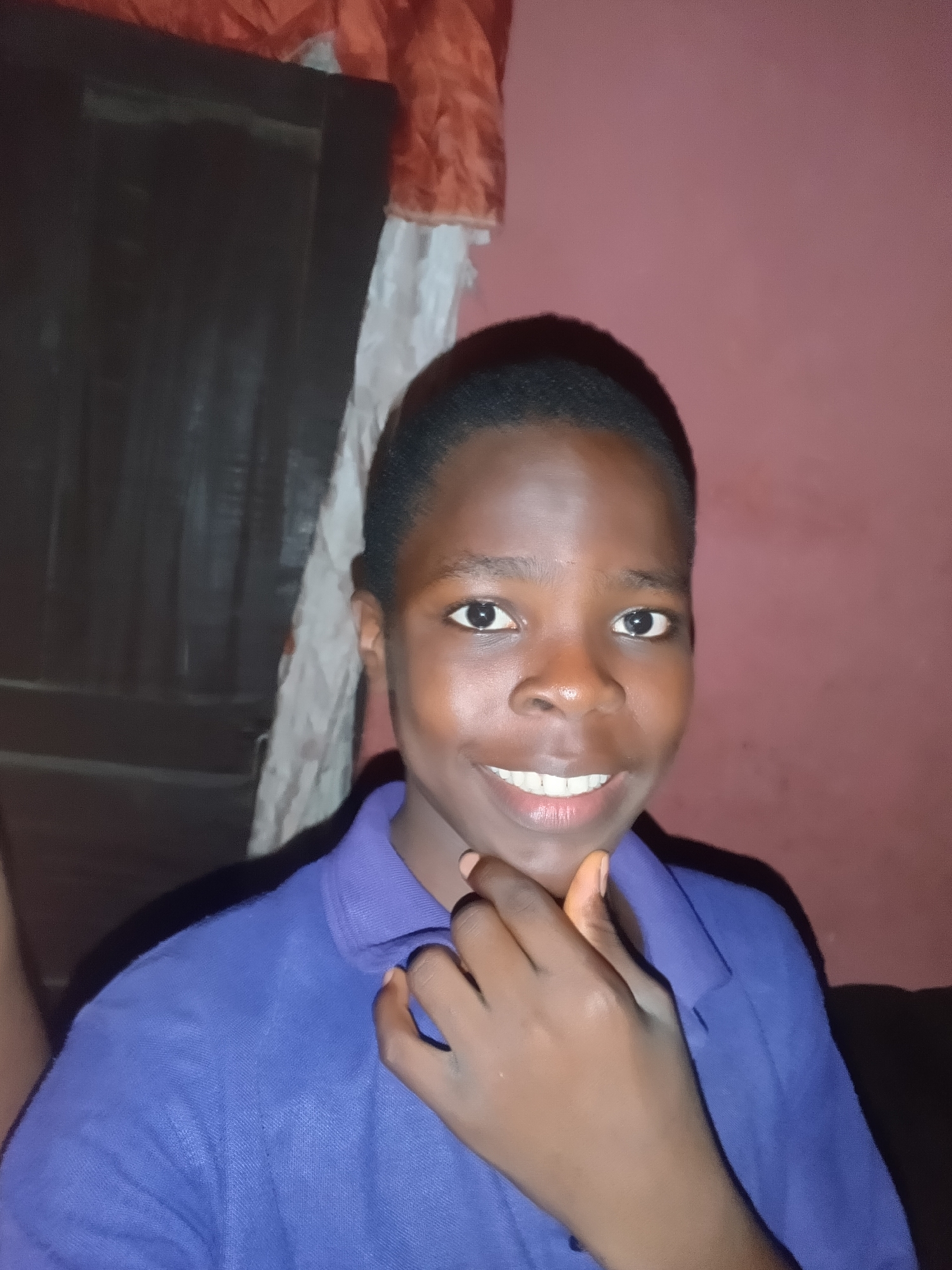 The image displays a head-and-shoulders portrait of a young individual with dark skin, positioned centrally and looking directly at the camera. The subject has short, dark, closely cropped hair. Their eyes are dark and wide open, exhibiting direct eye contact. A broad, open-mouthed smile reveals a row of white teeth. The individual is wearing a solid blue polo shirt with a visible collar and textured fabric. Their right hand (viewer's left) is raised, with fingers gently resting on their chin and jawline. The nails are short and unpolished.
The background indicates an indoor setting. To the viewer's left, a dark brown or black rectangular object, possibly a wooden door or piece of furniture with vertical paneling, is partially visible. Above and to the left of this object, a crumpled reddish-orange fabric or curtain hangs. A vertical strip of white or off-white wrinkled fabric is visible between the dark object and the wall. The remainder of the background, primarily to the right of the subject, consists of a plain wall painted a muted reddish-pink or light maroon color. Lighting appears to be consistent with indoor ambient conditions, casting subtle shadows. This scene is situated in the city of Onitsha, Nigeria.