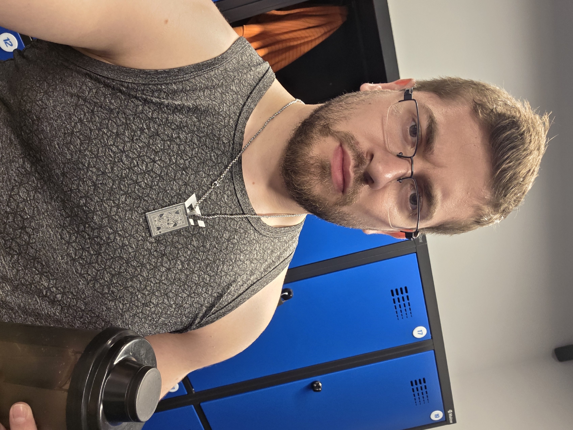A Caucasian male with a beard and glasses, wearing a gray tank top and a necklace with a playing card pendant, is holding a dark-colored shaker bottle. He is standing in front of a row of blue lockers, likely in a gym or sports facility. The location is not specified as Plovdiv, Bulgaria, and cannot be inferred from the image.