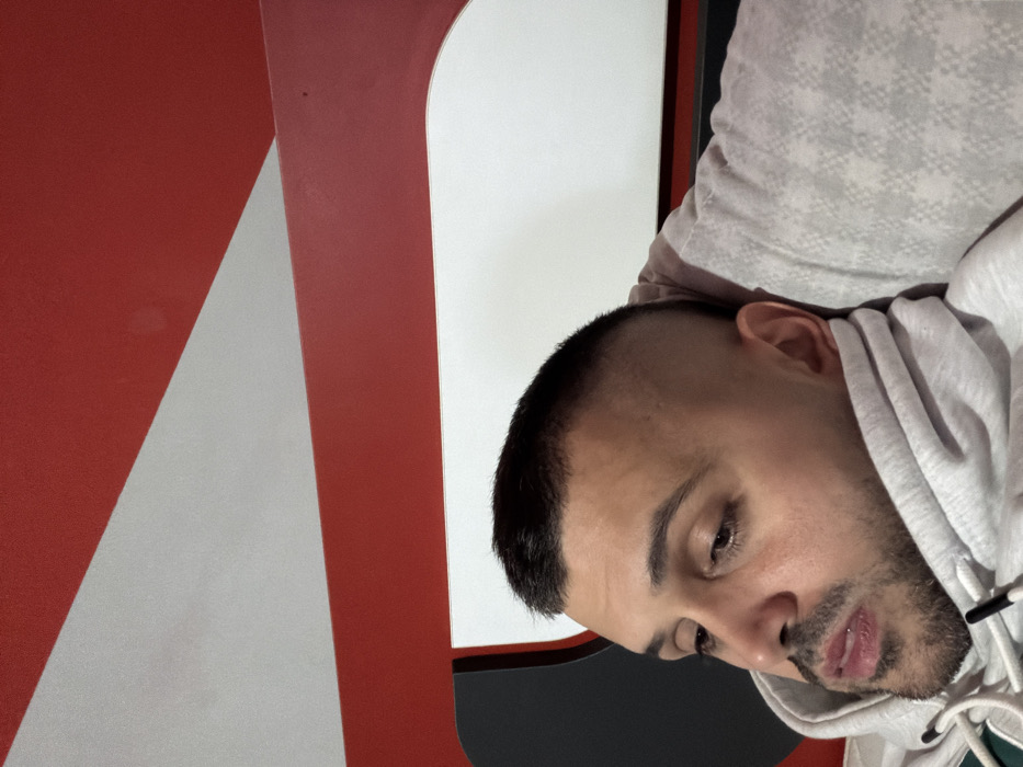 A male individual is depicted in a close-up, angled portrait, positioned in Opanets, Bulgaria. He has short, dark hair styled in a buzz cut with faded sides and dark stubble on his face. His skin tone is light to medium. He is wearing a light grey hooded sweatshirt, with the hood or collar drawn up around his neck and chin area. His head rests against a light-colored, possibly patterned, soft surface, likely a pillow or cushion, with a faint grid or diamond pattern visible. The subject's gaze is directed downwards and to the left of the viewer, with his mouth slightly ajar. The background consists of distinct geometric shapes: a prominent red horizontal band traverses the upper portion of the frame, bordered by white sections above and below. Diagonal red lines also intersect the white areas, creating angular patterns. A dark, curved element is visible on the far left edge of the frame.