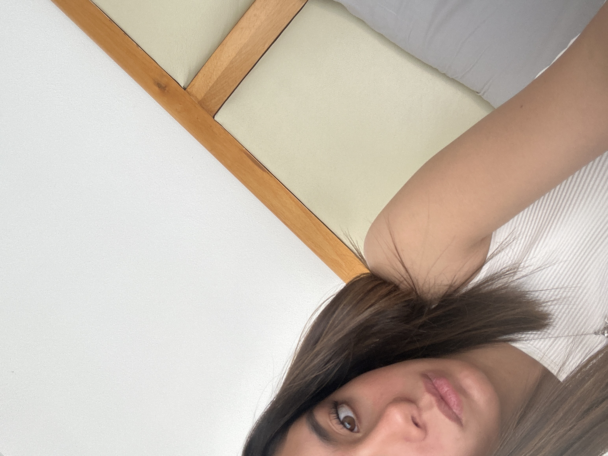 The image shows a close-up perspective of a person lying down, likely on a bed. The bed has a light-colored mattress and a wooden headboard. The person has brown hair and fair skin. They are wearing a white striped top. The background consists of a white wall. The location is unknown.