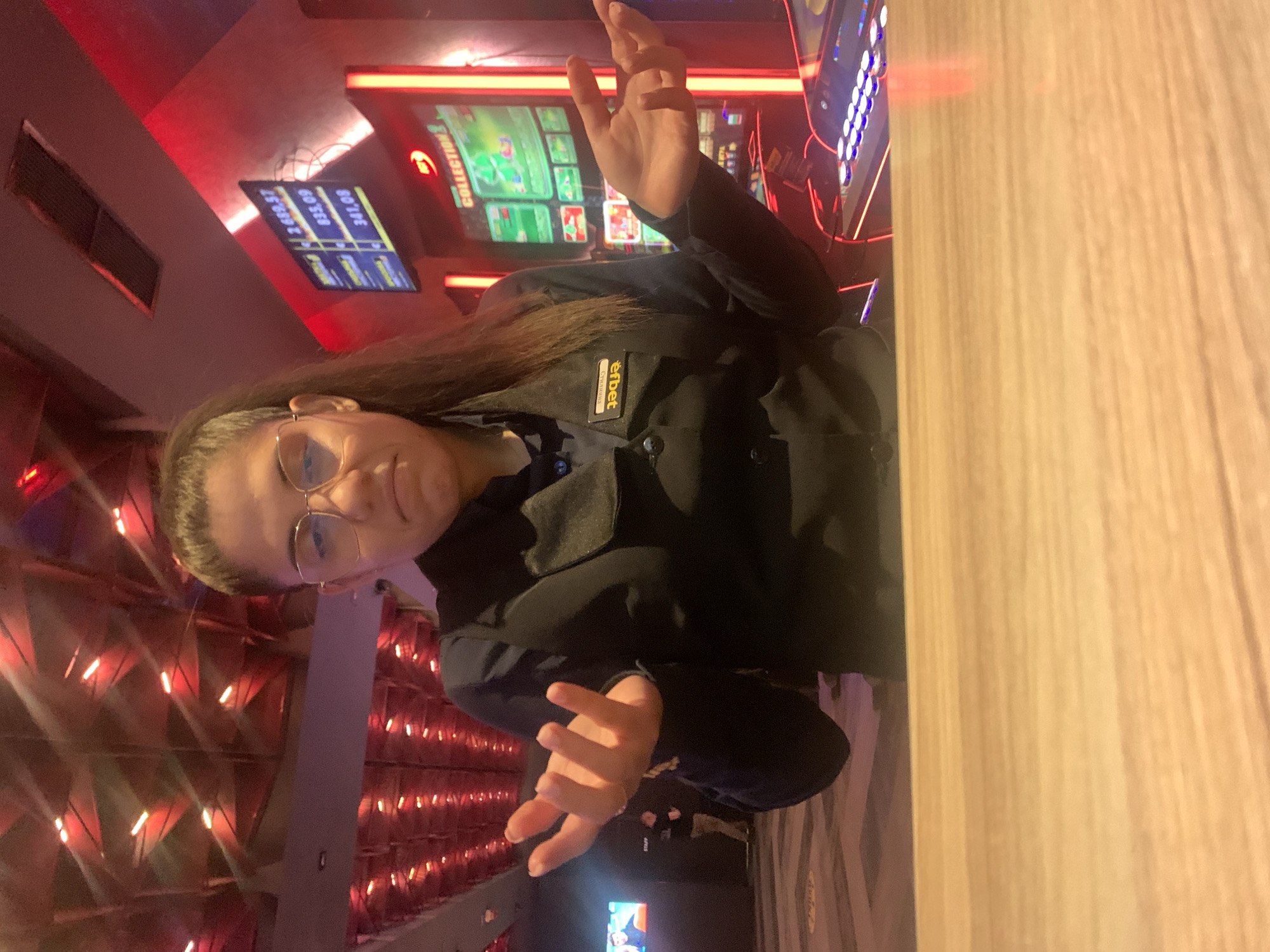 A young woman wearing glasses and a uniform with a name tag that reads "STAFF" in Bulgarian stands behind a wooden counter. She has her hands raised in a gesture of uncertainty or questioning. In the background, a brightly lit casino or gaming establishment is visible, with slot machines and digital displays showing numbers and graphics. The ceiling is illuminated with red lights. The overall scene suggests a customer service interaction in a casino setting in Burgas, Bulgaria.