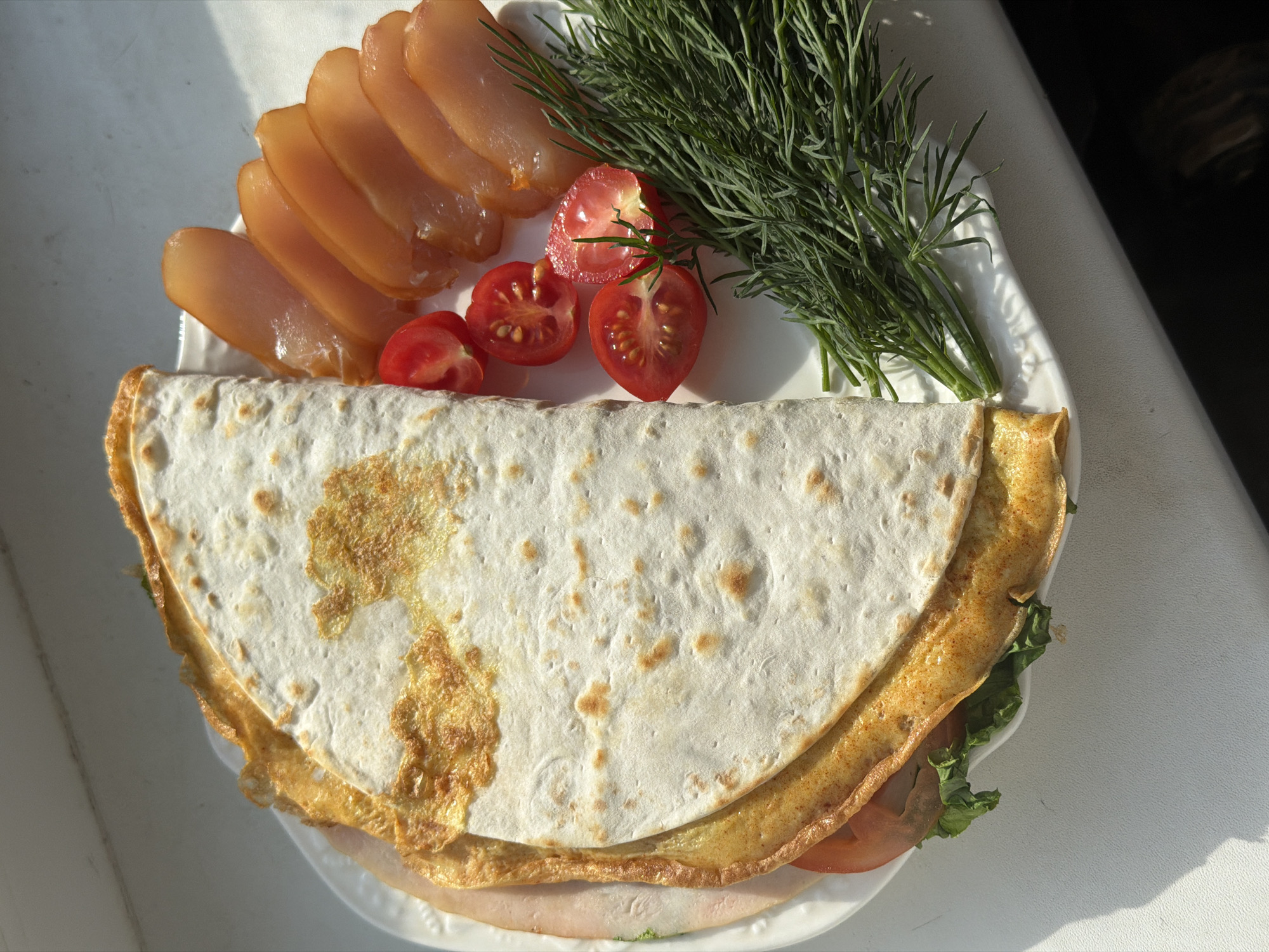 A photograph captures a white plate arranged with food items, likely taken in Perm, Russia. The plate displays slices of fish, halved cherry tomatoes, and a bunch of dill. A folded flatbread, seemingly filled with an egg and additional fillings, completes the dish. The arrangement sits on a white surface, possibly a table.