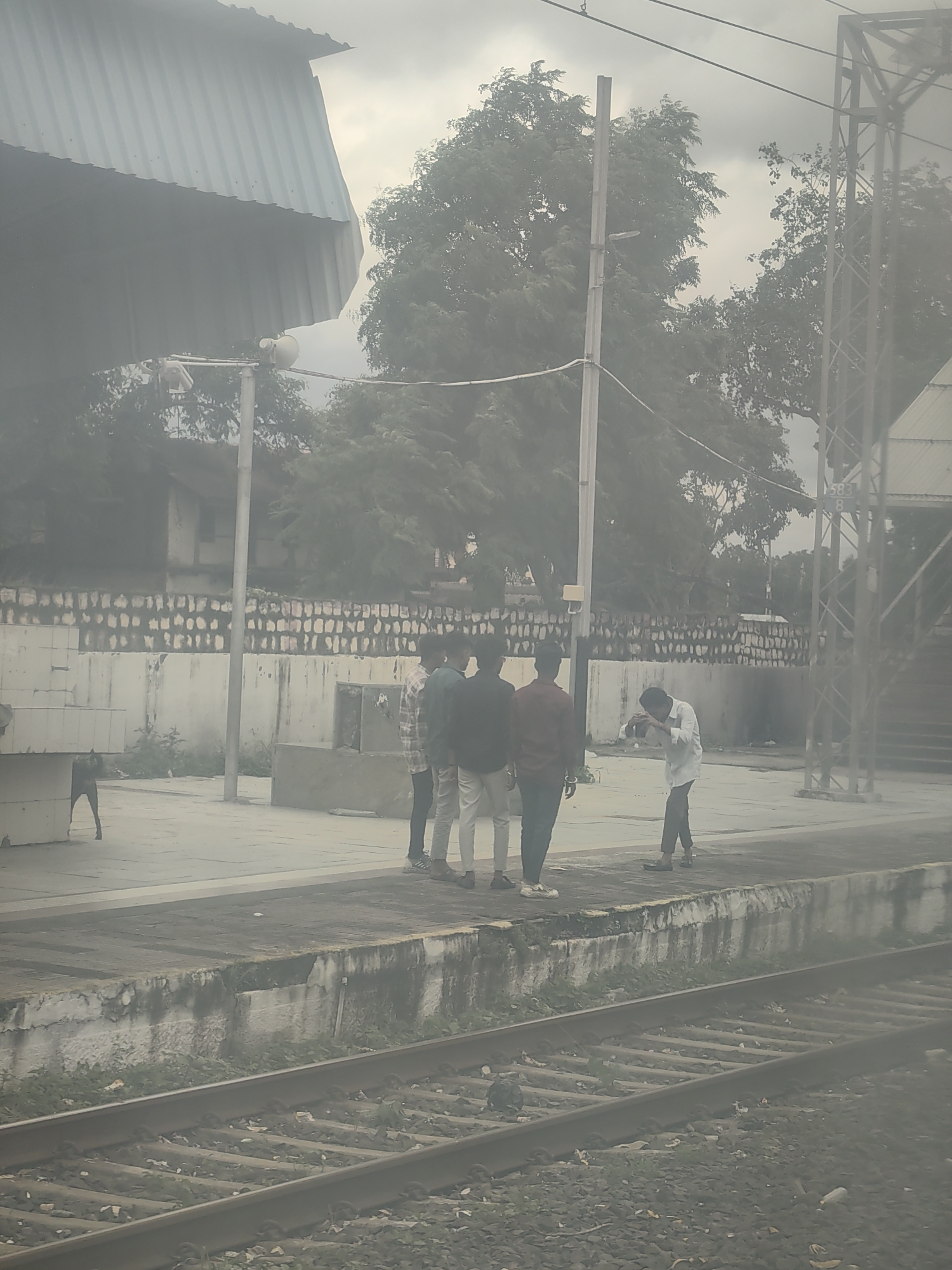 The image depicts a train station platform in Jawar, India, under an overcast sky. A group of four young men are standing on the platform. Three of them are positioned together, with one in a plaid shirt, another in a red shirt, and the third in a dark shirt, all facing towards the camera. A fourth man, wearing a white shirt and dark pants, is to their right, bending down as if to take a photograph or operate a device. A dog is visible on the platform in the background to the left. The platform itself shows signs of wear, with a concrete edge bordering the railway tracks. In the background, there is a low, stone-patterned wall and trees. A tall metal structure, likely part of the station's infrastructure, stands to the right of the frame. The image has a hazy or foggy quality, obscuring some finer details.