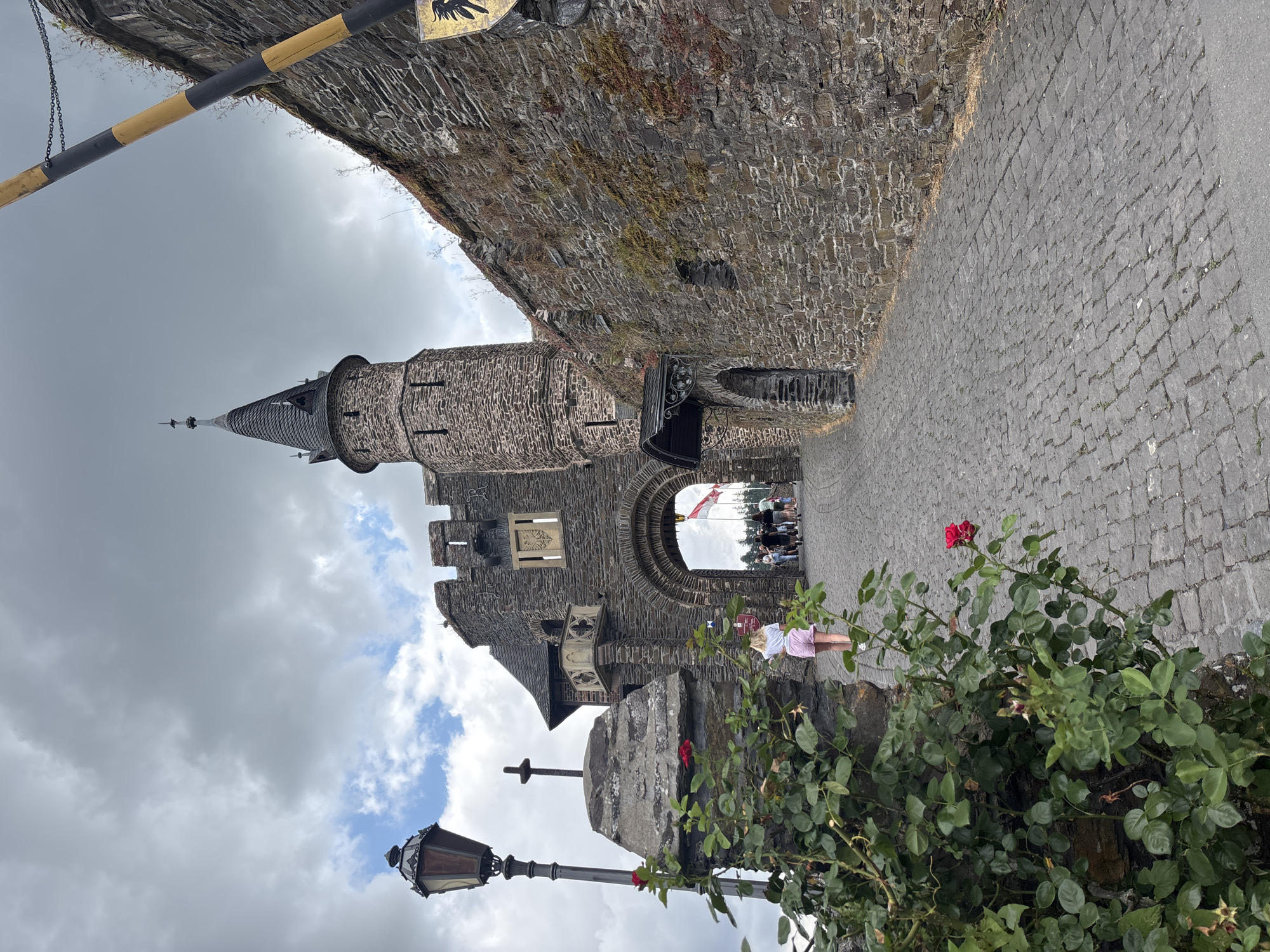 The image captures an upward view of a cobblestone path leading through a stone gateway to a castle, possibly Reichsburg Cochem, in Cochem, Germany. In the foreground on the left, a lush green rose bush with a single red rose is in bloom, positioned against a stone pillar topped with a cross. A vintage lamp post stands beside the pillar.
The stone gateway features a large arched entrance and a circular tower with a pointed roof, characteristic of medieval architecture. The walls are constructed from rough-hewn stones, some adorned with small windows and decorative elements. A yellow and black striped barrier is visible on the right side of the image, suggesting an entrance control point.
Several people, appearing to be tourists, are walking through the archway into the castle grounds. A young woman in a white shirt and pink shorts is walking away from the camera on the cobblestone path. The sky above is overcast with some patches of blue. The overall impression is a historic and somewhat imposing, yet inviting, scene of a German castle.
