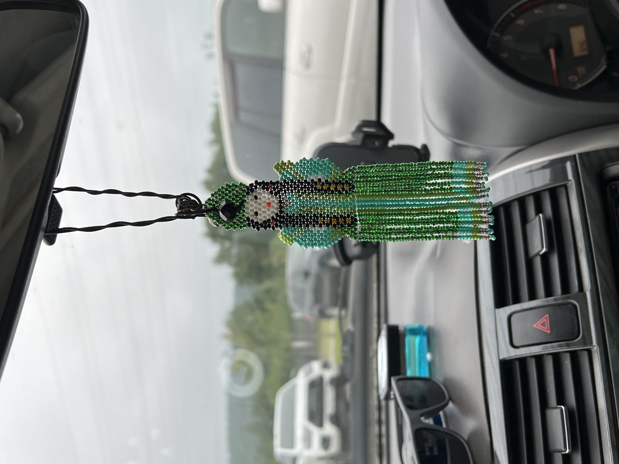 The image shows the interior of a car, looking out the windshield. In the foreground, on the dashboard, is a beaded doll hanging from the rearview mirror. The doll is green and blue, with a beaded headdress and fringe. To the left of the doll are a pair of sunglasses and a blue air freshener. In the background, several cars are parked. The sky is overcast. The scene is consistent with a location in the city of Yuzhno-Sakhalinsk, Russia, given the weather and the type of vehicles visible.