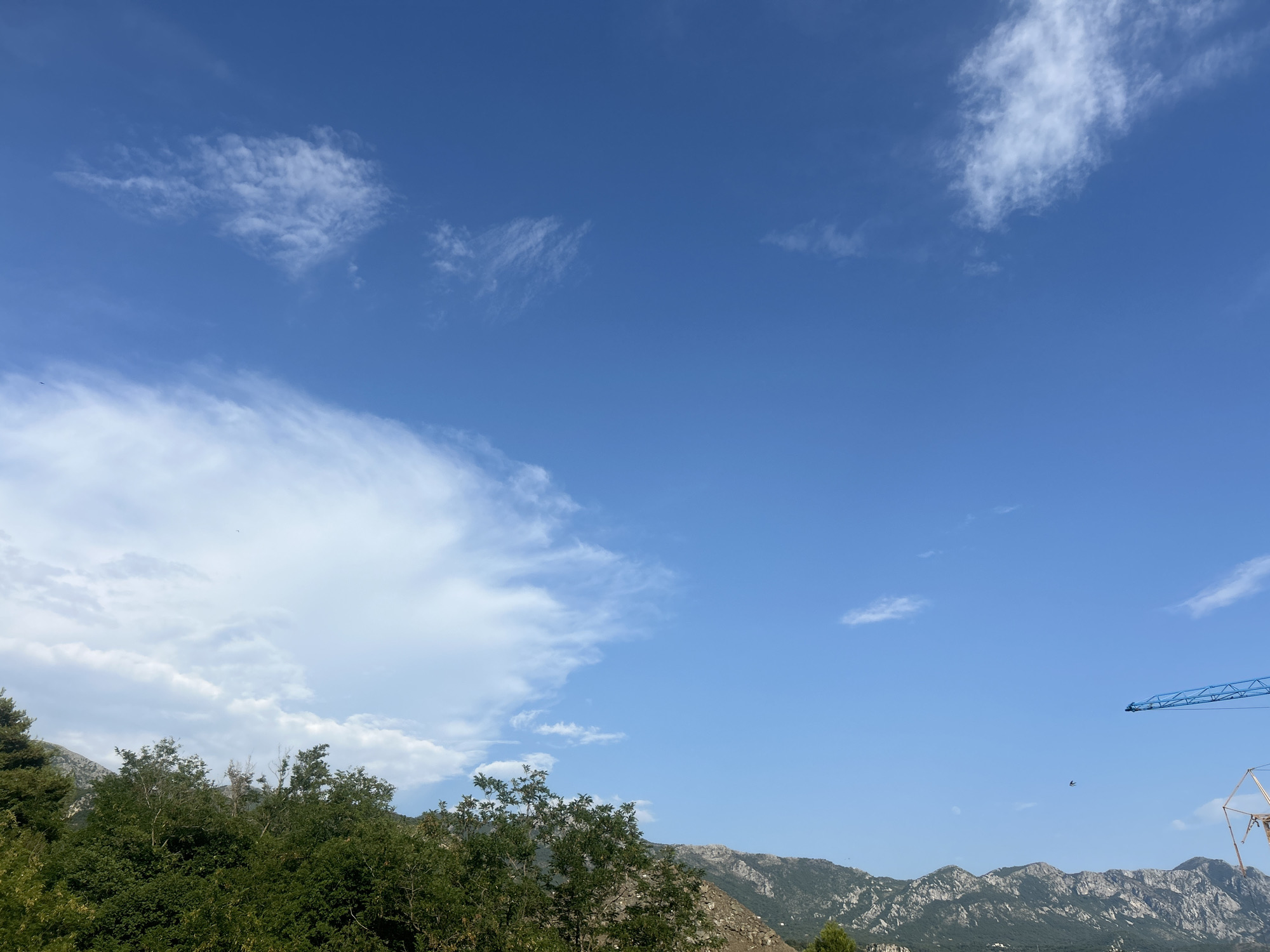 This is a landscape photograph taken outdoors on a clear day in Boreti, Montenegro. The scene is dominated by a vast blue sky with scattered white clouds. In the lower portion of the image, lush green trees and foliage create a dense canopy. Beyond the trees, a range of rugged, mountainous terrain stretches across the horizon, with some areas appearing rocky and barren while others are covered in vegetation. A construction crane, painted blue, is visible in the upper right corner of the image, suggesting ongoing development in the area. A small bird is also visible in flight against the bright sky. The lighting indicates it is daytime, likely late morning or early afternoon, with bright sunlight casting clear shadows. The overall atmosphere is serene and natural, with a hint of human intervention from the construction equipment. There is no visible text in the image.