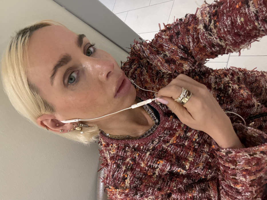 A fair-skinned person with short, blonde hair faces the camera, wearing earbuds. The individual has visible freckles and rings adorn their fingers. They wear a multi-colored, tweed-like sweater or jacket in shades of red, orange, and brown. A wall is visible behind the person, suggesting an indoor setting. The overall composition is a casual selfie.