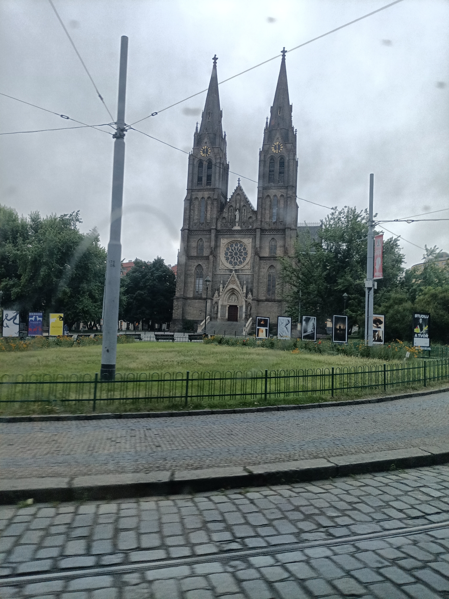 The image shows the Basilica of Saint Peter and Saint Paul located in Vyšehrad, Prague, Czechia. The basilica, a neo-Gothic Roman Catholic minor basilica, is characterized by its two prominent spires. In front of the basilica, a green lawn is bordered by a dark green metal fence. Several large framed displays of artwork are situated on the lawn, with some exhibiting posters or advertisements. Behind the lawn, the basilica stands tall, its facade featuring a large rose window above the main entrance and statues. Tall green trees are visible on either side of the basilica and behind it, partially obscuring some of the building. Overhead, power lines for what appears to be a tram system crisscross the sky, with a tall gray pole supporting them in the foreground. The ground in front of the lawn is paved with cobblestones, and tram tracks are visible embedded in the pavement. The sky is overcast and gray.