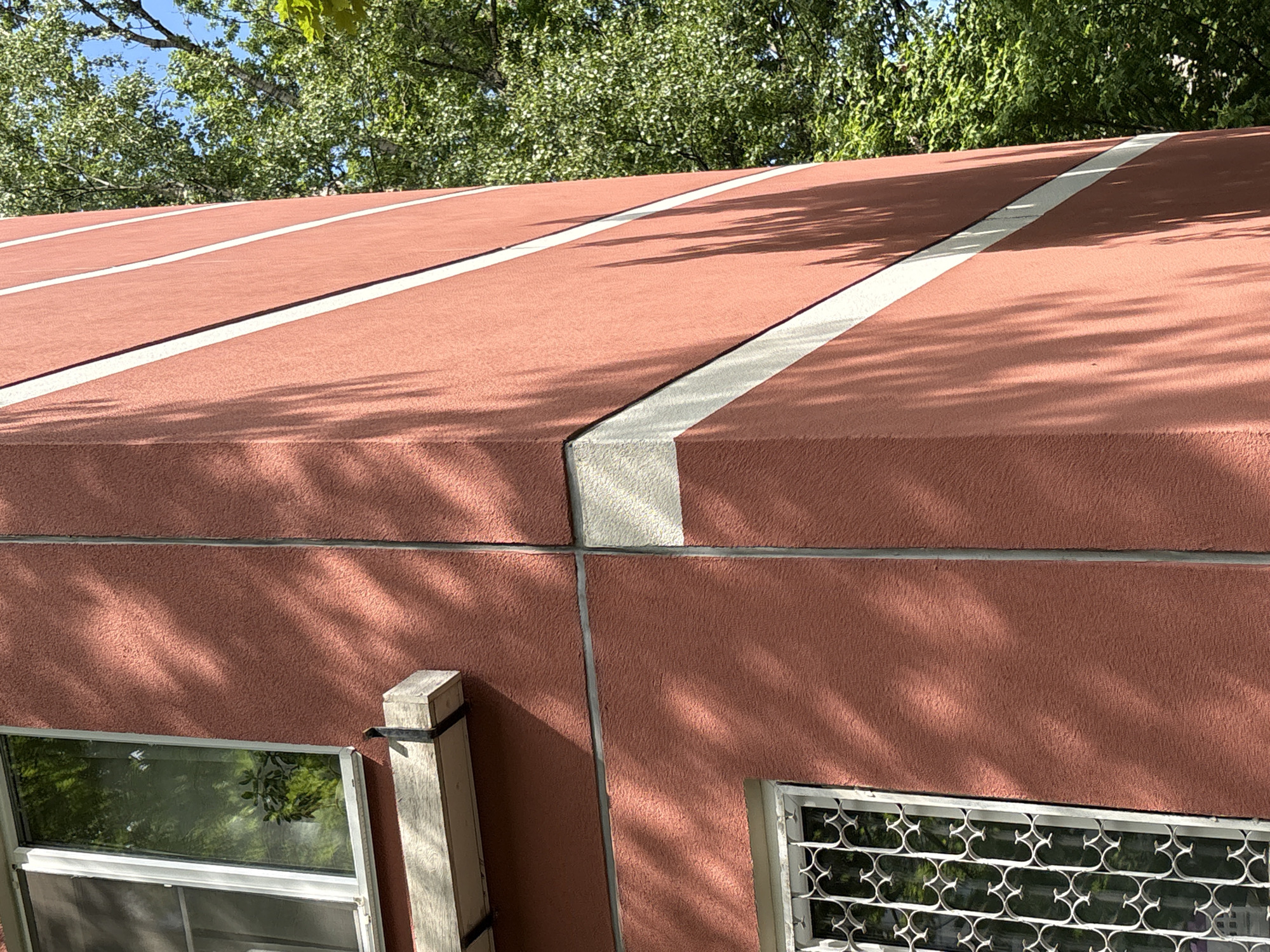 The image displays an exterior view of a building, focusing on its reddish-brown roof and upper wall section, set against a backdrop of vibrant green trees and a blue sky. The roof features a granular or stucco-like texture, with prominent white or light gray stripes running diagonally across its surface, resembling architectural seams or decorative elements.
Below the roof, the building's wall continues with the same reddish-brown, textured finish, marked by a horizontal gray division line. Sunlight casts intricate, dappled shadows of tree leaves and branches across both the roof and the wall, indicating a bright, sunny day.
At the bottom of the frame, two windows are partially visible. The window on the left has clear glass, reflecting the surrounding foliage. The window on the right is protected by an ornate white metal security grate featuring a recurring, stylized pattern. A weathered wooden post, possibly a structural support, is seen on the lower-left, secured to the wall. No people or specific activities are depicted, and no text is visible. The scene captures architectural details under natural daylight.
