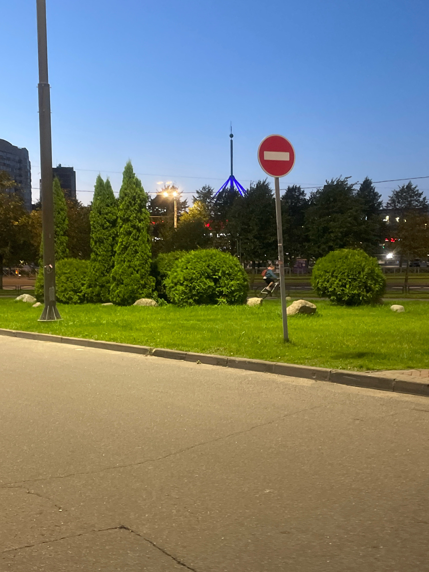 The image depicts a street scene at dusk in Saint Petersburg, Russia. A paved road occupies the foreground, with a sidewalk and a grassy median strip beyond. The median is adorned with several conical evergreen trees and rounded bushes, interspersed with large rocks. A traffic sign, a red circle with a horizontal white bar, is prominently displayed on a pole in the median. In the background, several apartment buildings and a tall, illuminated tower with a blue light structure at its base are visible. A person on a bicycle is seen riding through the median. The sky is a clear, deep blue, indicating early evening. Streetlights are also illuminated in the distance.