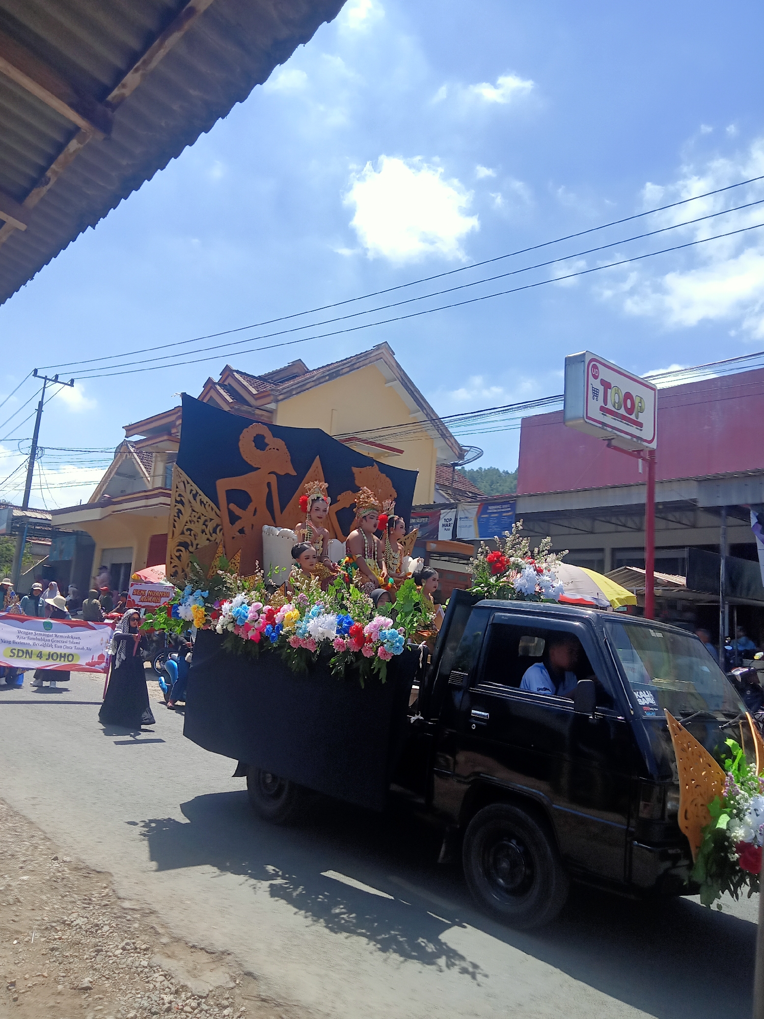 The image depicts a street parade in Pule, Indonesia, on a sunny day with a bright blue sky dotted with clouds. In the foreground, a black pickup truck is adorned with colorful flowers and features a backdrop of a traditional Indonesian wayang kulit puppet. Two individuals, dressed in traditional attire, are seated on the truck's decorated platform. A banner held by two people in the middle ground reads "DENGAN SEMANGAT KEMERDEKAAN, KITA TUMBUHKAN GENERASI MUDA YANG BERIMAN, BERAKHLAK DAN CINTA TANAH AIR SD N 4 JOHO". The street is paved and appears to be in a somewhat rural or suburban area, with buildings and power lines visible in the background. Several other individuals are present on the sides of the street, observing the parade. The overall atmosphere suggests a celebratory event.