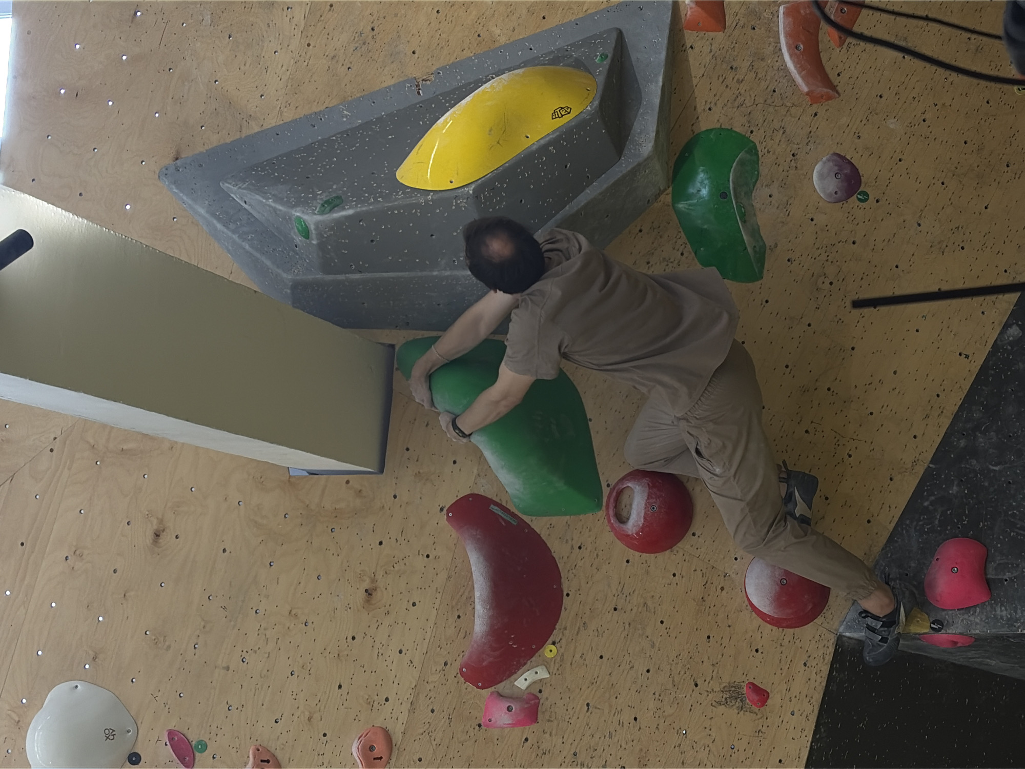 A man is rock climbing on an indoor climbing wall. He is wearing a light brown t-shirt and beige pants, with black climbing shoes. He is reaching for a green climbing hold. The wall is made of wooden panels and has various colorful climbing holds attached to it. The image was taken in Moscow, Russia.