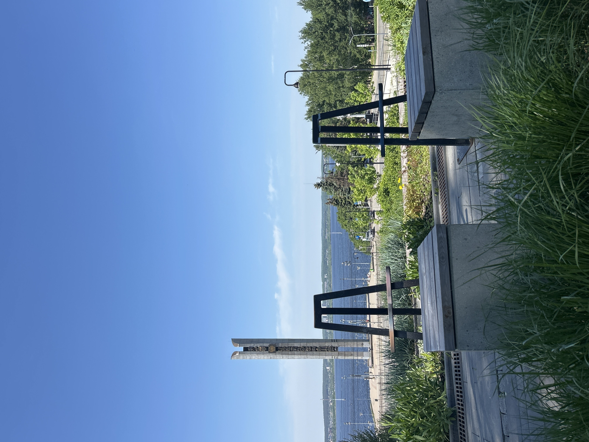 The image shows a view from a park in Izhevsk, Russia. In the foreground, there are concrete benches with metal frames, surrounded by greenery. Beyond the benches, there is a monument with vertical inscriptions, and a body of water (likely the Izhevsk reservoir). Along the reservoir, there is a paved walkway with streetlights. The background features a clear blue sky with some wispy clouds.