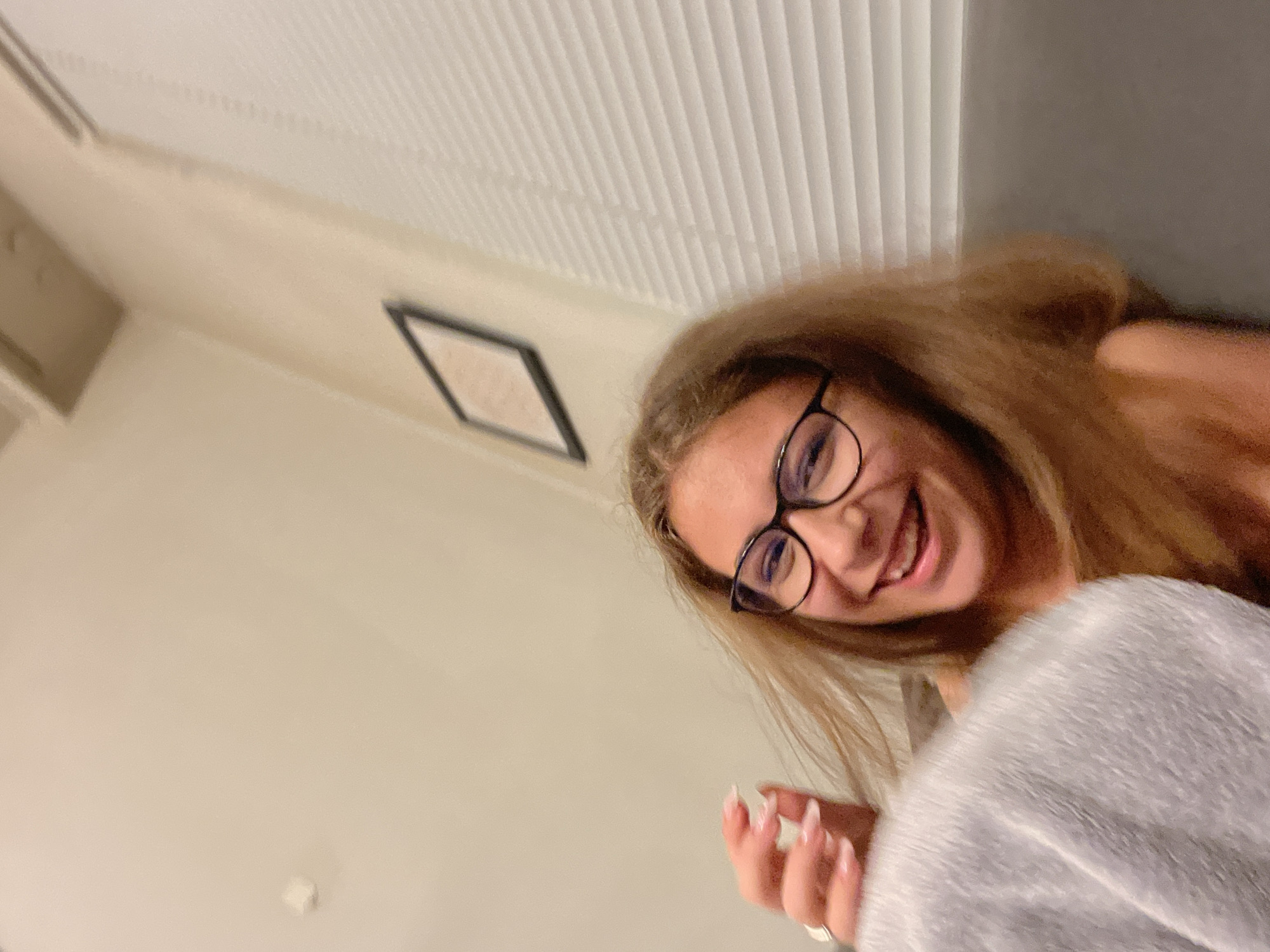 A young woman wearing glasses smiles into the camera. She is indoors, possibly in a bedroom, as indicated by the blinds on the ceiling and a framed picture on the wall. She is wearing a fuzzy grey robe or jacket. The location is likely Plovdiv, Bulgaria, but this is unconfirmed within the image itself. There are no clear indicators of a city location.