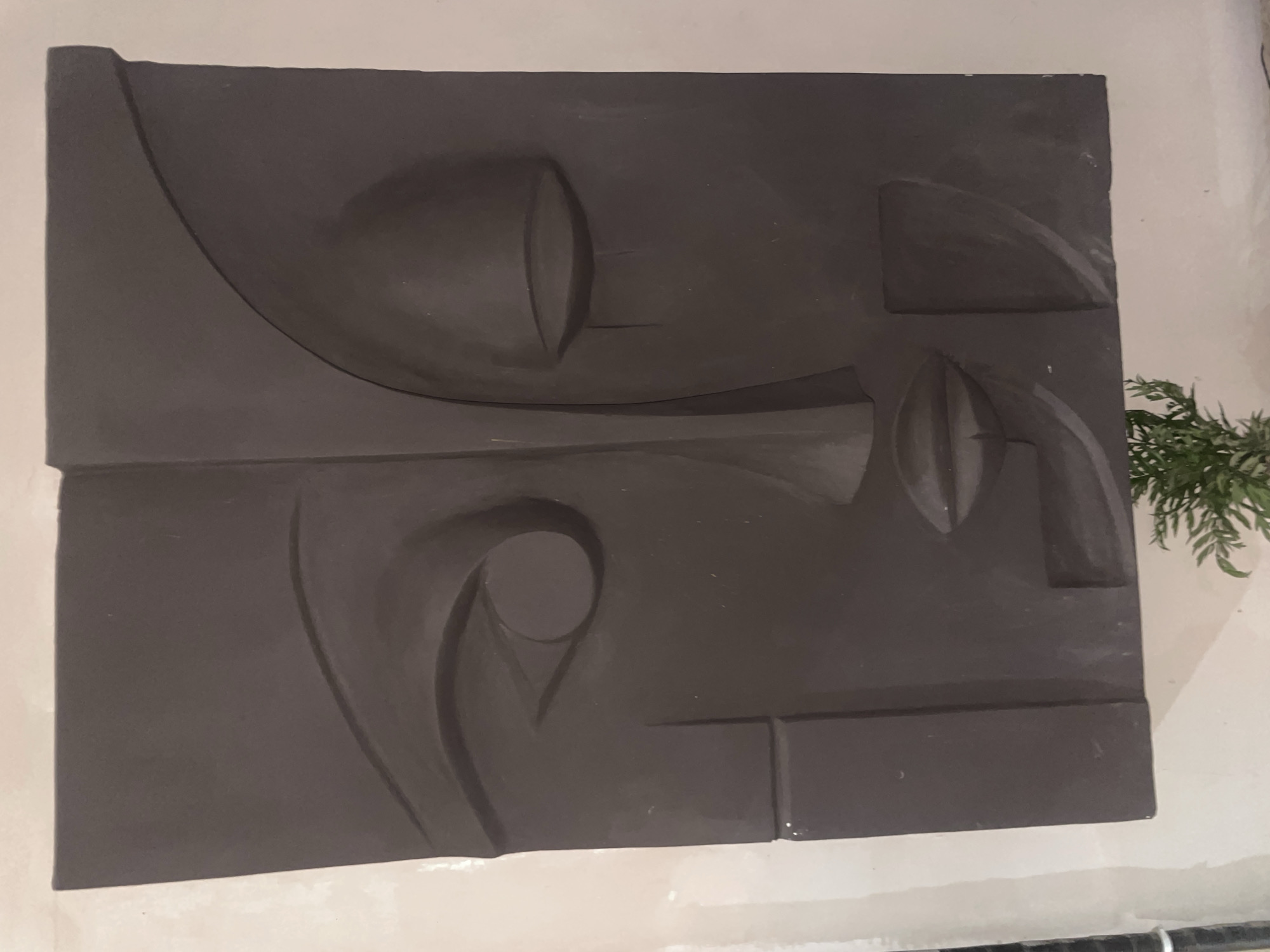 This image captures a close-up view of a dark gray, abstract relief sculpture of a face mounted on a light-colored wall. The sculpture is a stylized representation, with geometric shapes defining the features. The left side of the face shows an open eye with a circular pupil, while the right side features a closed eyelid with a tear-like line extending downwards. The lips are rendered as a horizontal, slightly curved shape. The background wall appears textured and possibly under construction, with visible seams and imperfections. At the bottom of the frame, a small section of green foliage, likely from a potted plant, is visible. The overall lighting is soft, casting subtle shadows that emphasize the dimensionality of the sculpture. The scene is devoid of people, focusing solely on the artwork and its immediate surroundings. The setting suggests an indoor environment, possibly an art gallery, studio, or a home decorated with art.