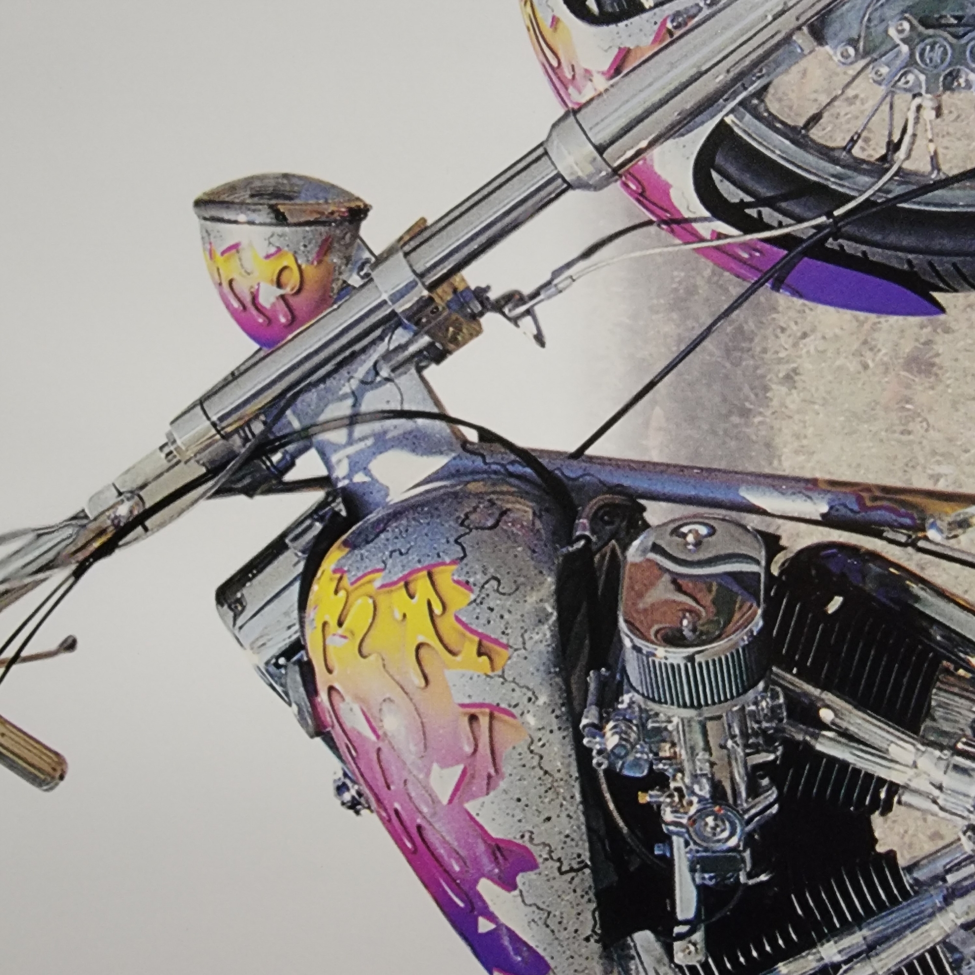 A close-up, eye-level, outdoor, slightly low-angle, medium shot captures the front of a custom motorcycle. The motorcycle's fuel tank is painted with a vibrant flame design in shades of pink, purple, and yellow, against a textured, metallic gray background. The front headlight, also decorated with a flame design, is visible on the right side of the image. Chrome accents are present throughout the handlebars, front forks, and the engine, which features a prominent air cleaner. The front wheel and part of the purple-painted front fender are also visible. The background consists of a softly blurred, outdoor environment with a muted, beige-colored ground and a light gray sky, suggesting an open, possibly rural setting rather than an urban one. There are no people or signs of a city present in the image.